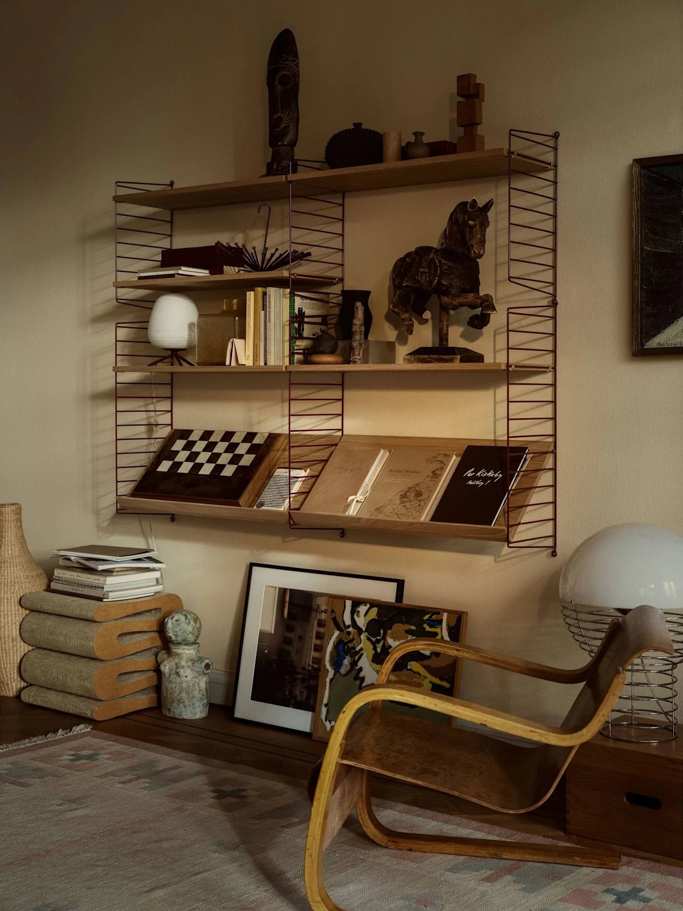 Et hyggeligt værelse med et reolsystem fyldt med bøger, kunst og indretning, ved siden af en træ lænestol og en stor hvid lampe.