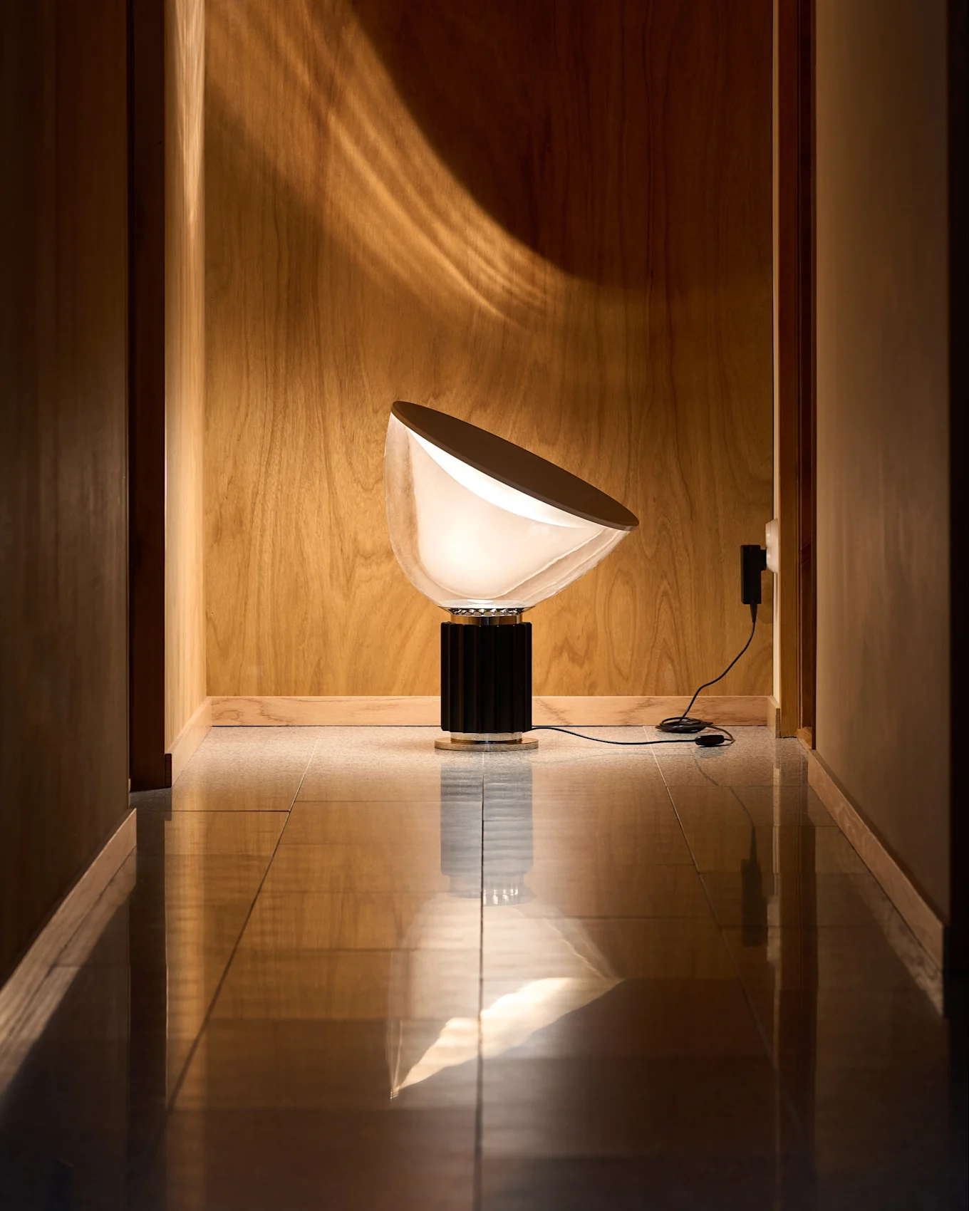 Moderne lampe med halvrund, hvid skærm og sort rillet fod i træbeklædt gang med reflekterende gulv. Lyset danner kurvet mønster.
