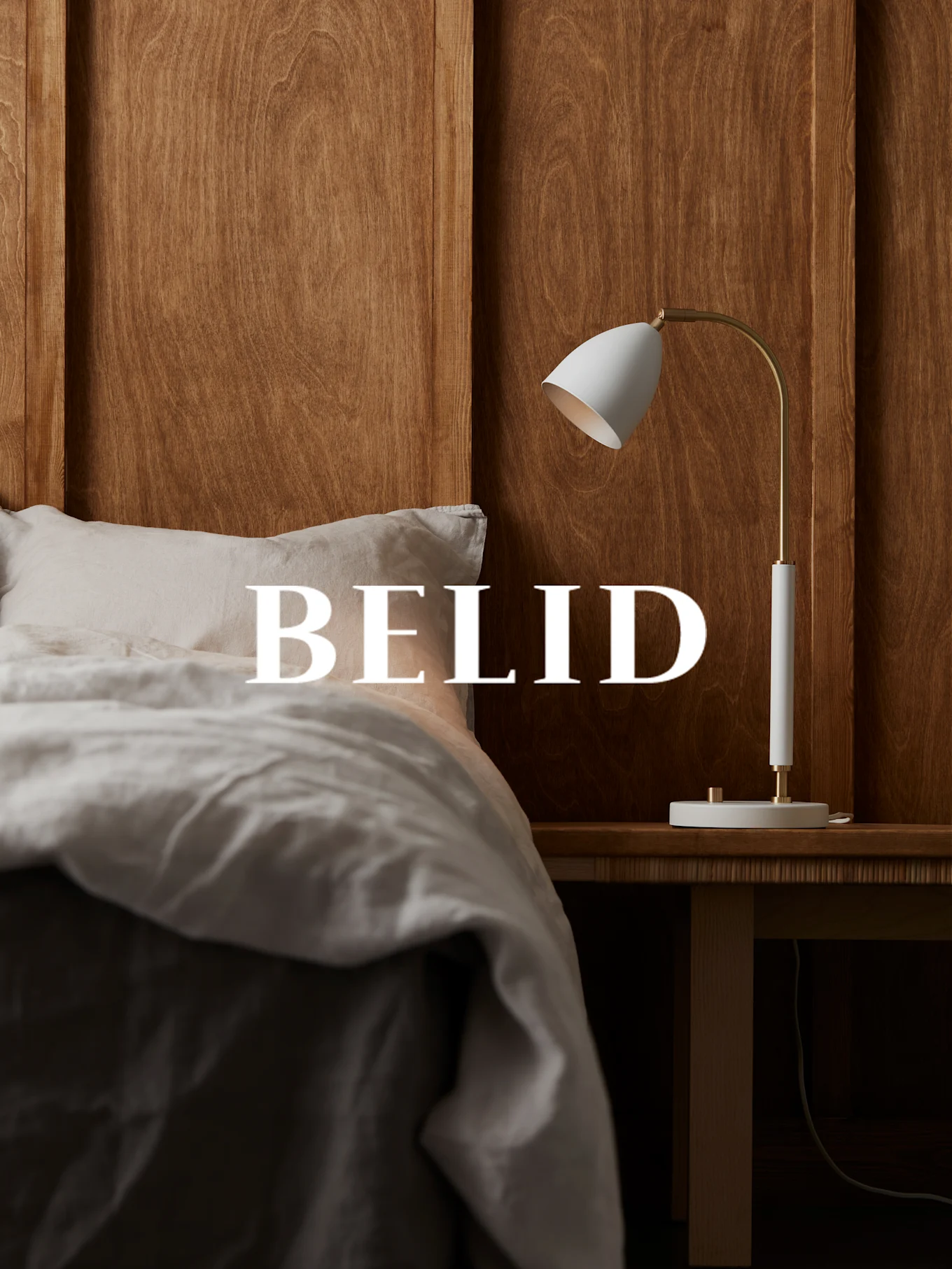 Hvid og messingfarvet lampe på et sengebord af træ, med teksten 'BELID' over en seng med linned sengetøj.