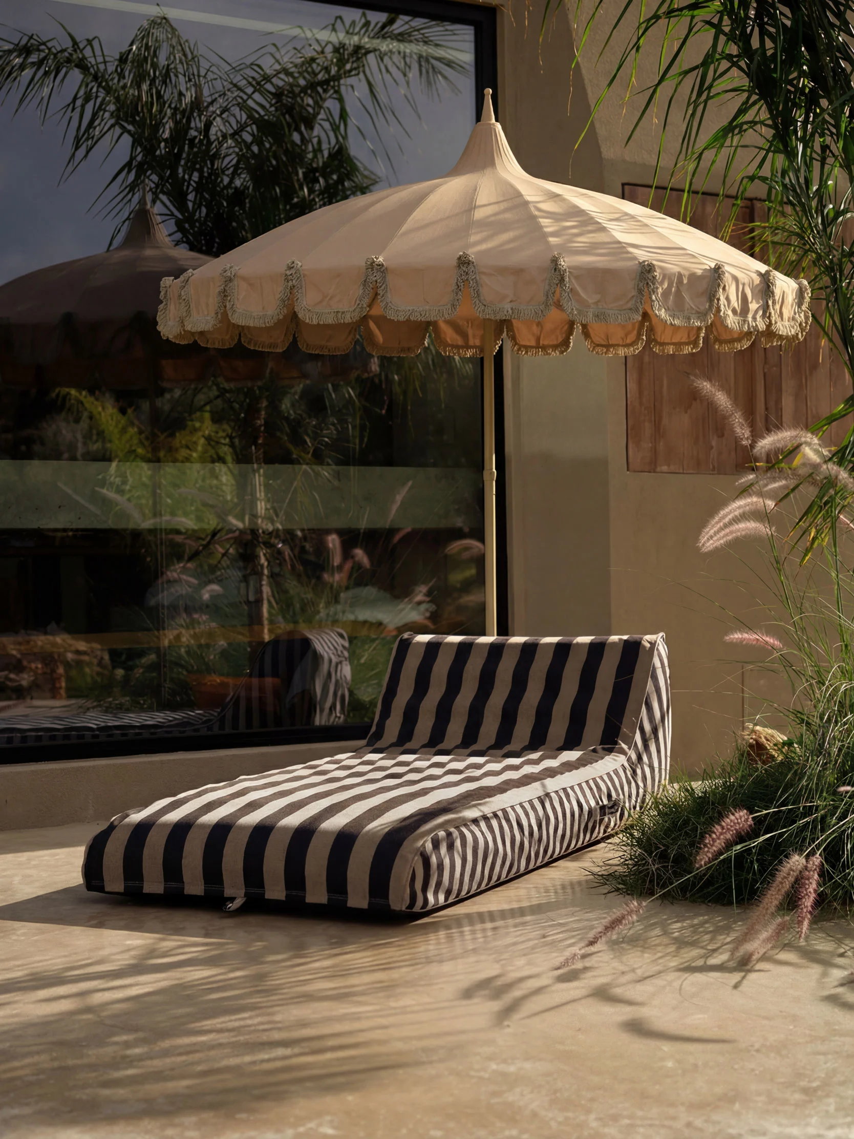 Udendørs terrasse med beige frynset parasol og stribet liggehynde på lyst gulv, omgivet af frodige planter.