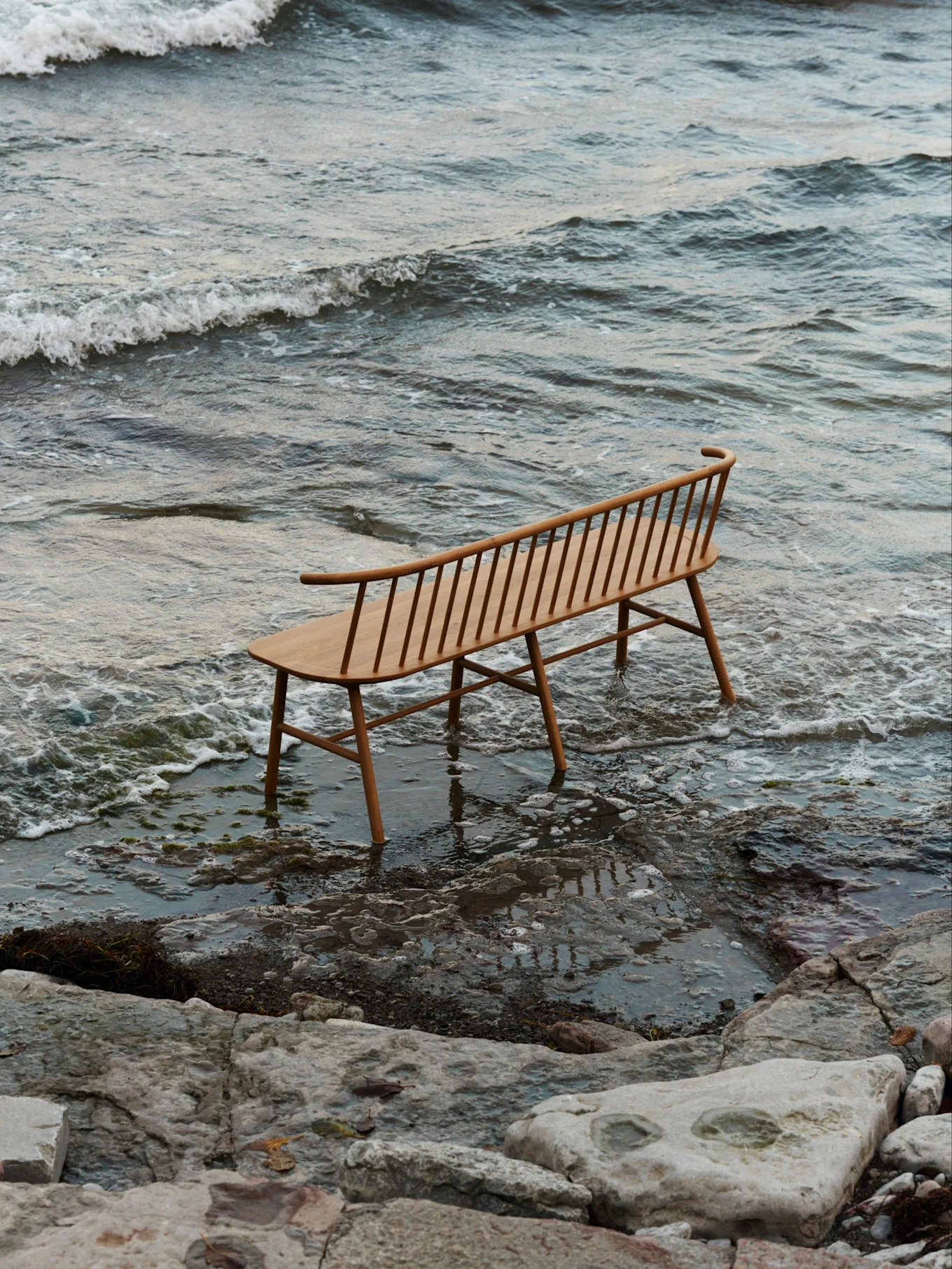 En lys træbænk med lameller og buet ryglæn står i lavt vand på en stenstrand med blide bølger.