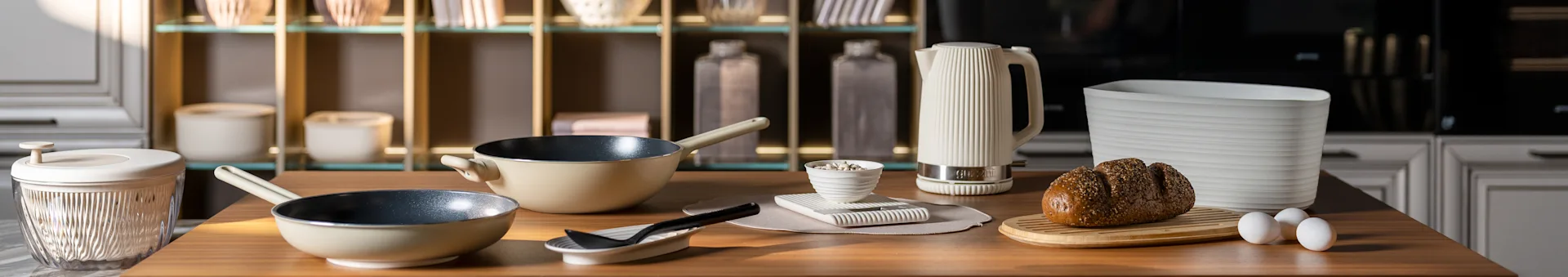 Køkkenbord med beige stegepander, en hvid riflet elkedel, et frøfyldt brød på et bræt og æg. Moderne køkkentilbehør.