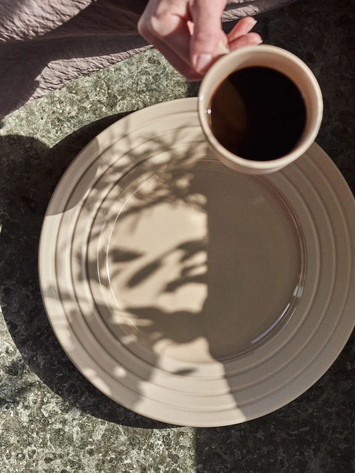 Hånd holder kaffekop over beige tallerken med bladskygger på et plettet bord.