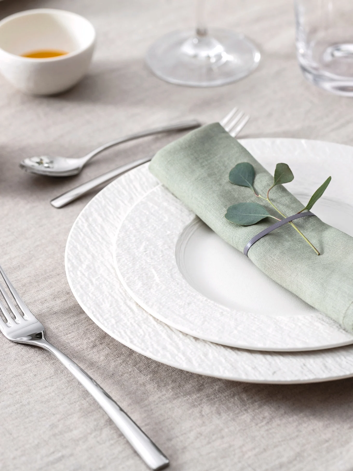 Elegant borddækning med lysegrøn linnedserviet, eukalyptusblade, hvide teksturerede tallerkener og bestik på en beige dug.