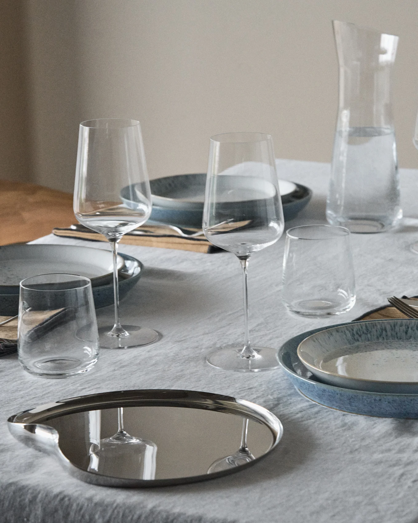Et dækket bord med blå og plettede tallerkener, vinglas, vandglas, en vandkaraffel og en reflekterende oval bakke.