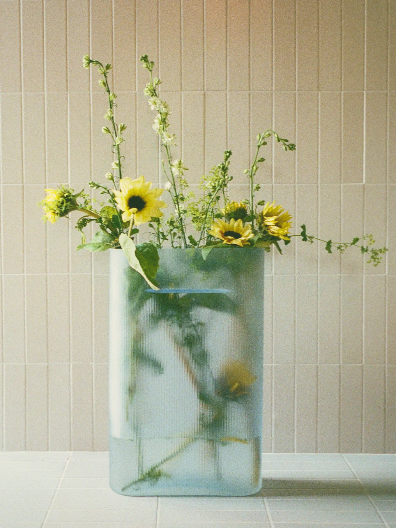 Buketter af gule solsikker og grønne spidse blomster i en klar, riflet rektangulær vase på en flisebelagt overflade.