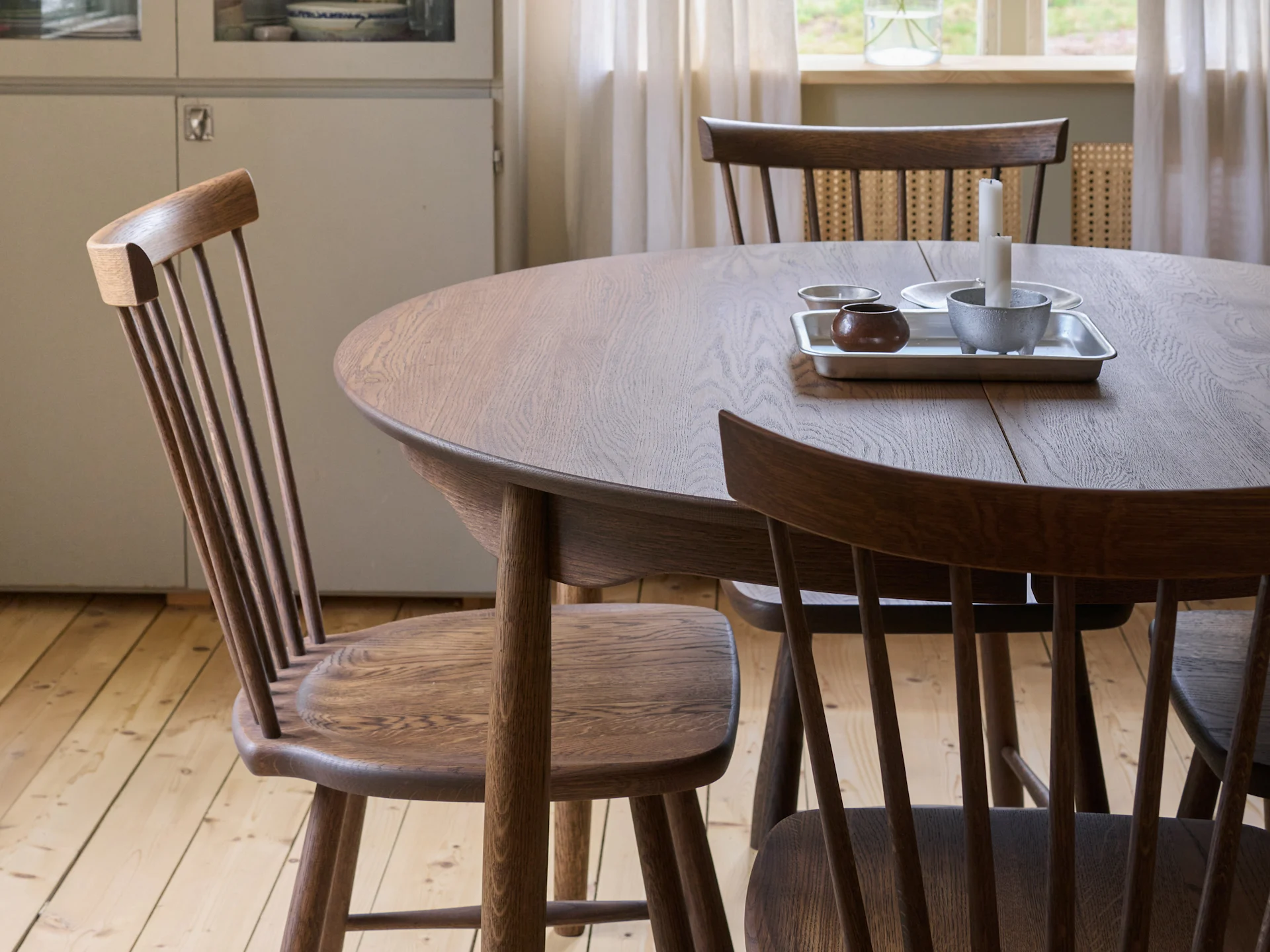 Et rundt spisebord i træ med tre stole med spindelryg, en bakke med lys og en skål, og et trægulv.