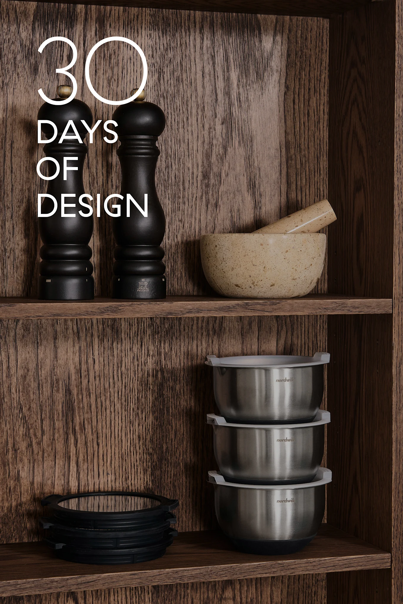 Træhylder med køkkenudstyr: sorte kværne, morter og støder, stålskåle, silikoneringe. Tekst: 30 DAYS OF DESIGN.