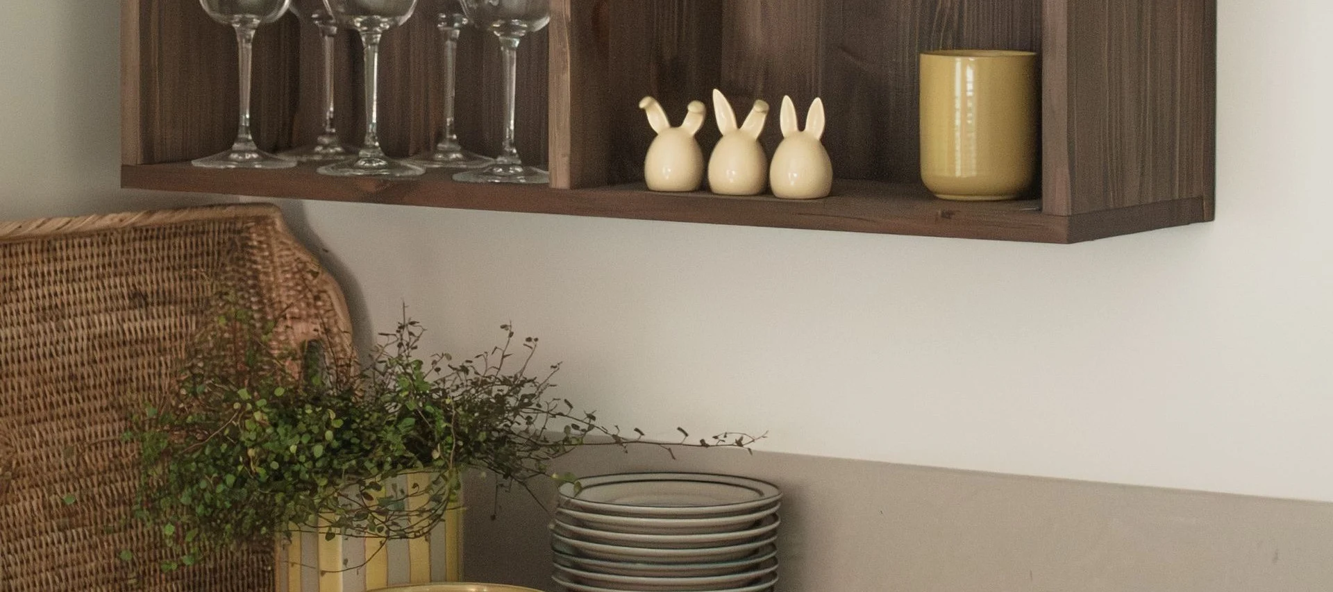 En træhylde med glas, tre kaninfigurer og et gult krus, med en fletkurv og planter nedenunder.