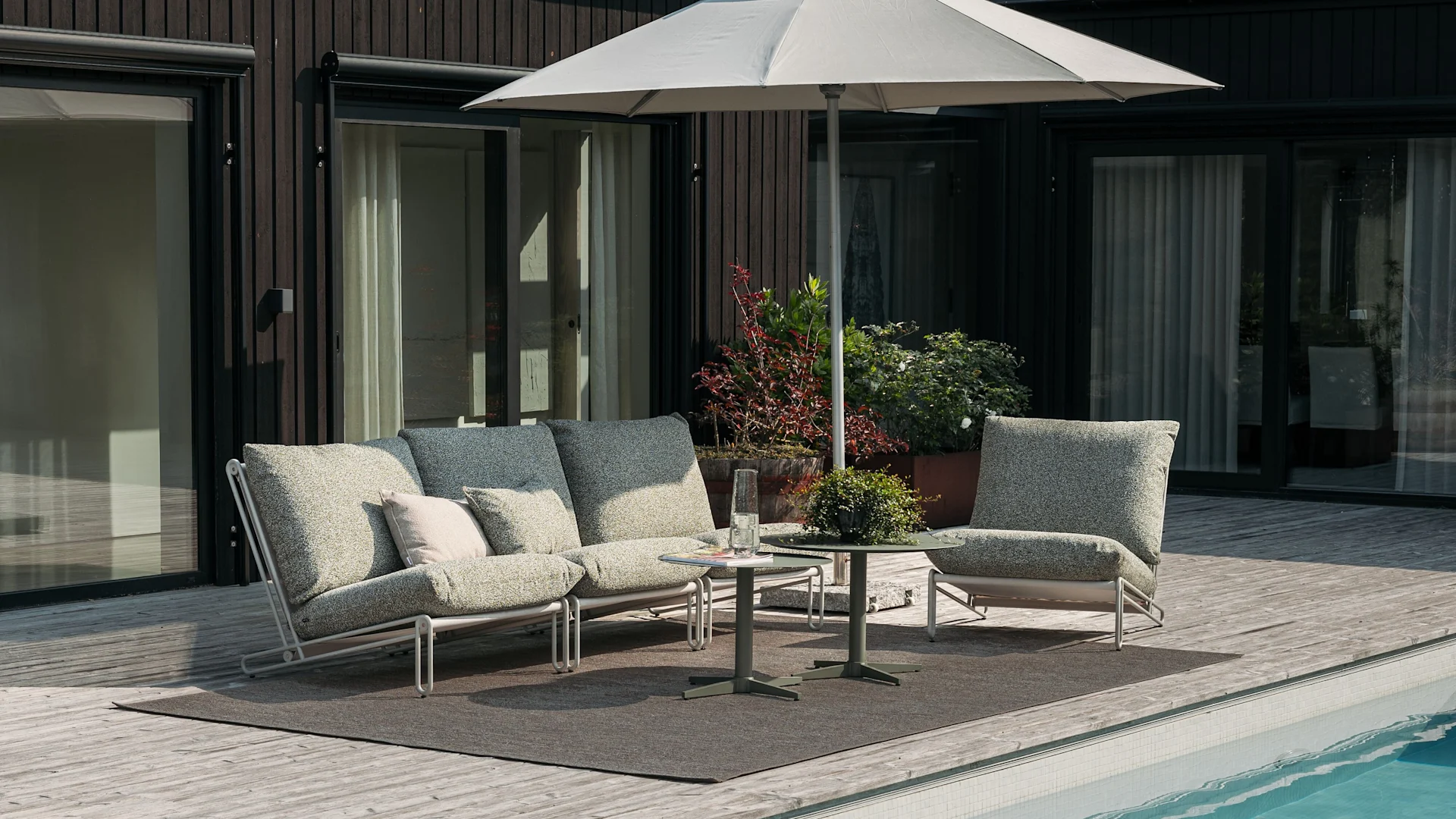 Udendørs terrasseområde med en lysegrå to-personers sofa, matchende lænestol og et rundt sofabord i glas på et gråt tæppe, ved siden af en pool og et mørkt træhus.