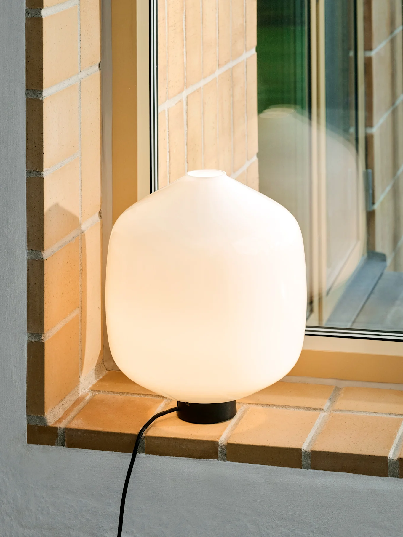 En lysende, afrundet hvid bordlampe med sort fod og ledning på en muret vindueskarm.