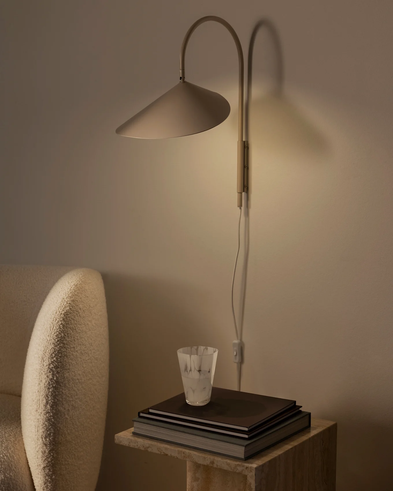 Varmt belyst væglampe med konisk skærm over en stak bøger og et tekstureret glas på et stenbord, ved siden af en beige lænestol.