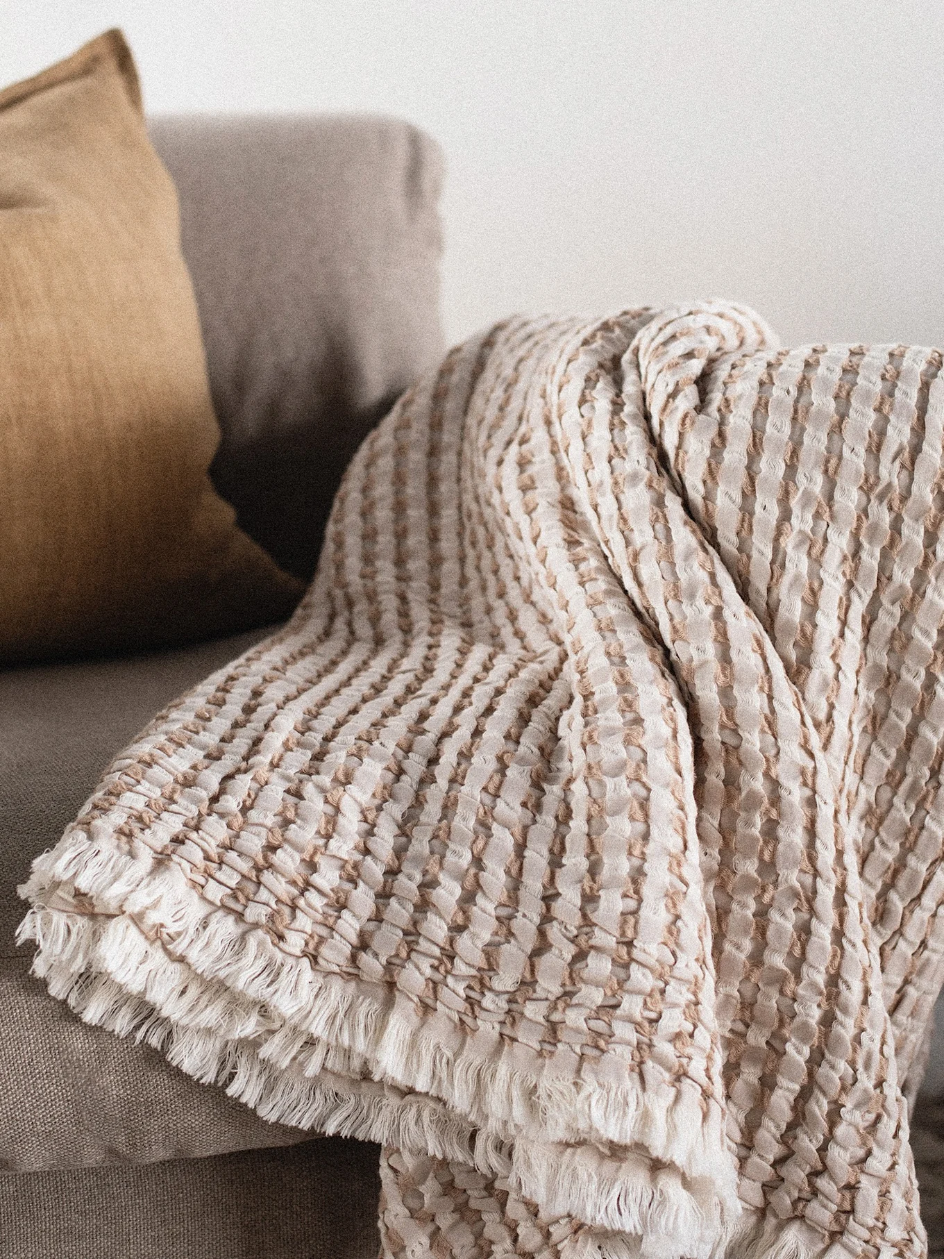 Tekstureret lys beige og brun stribet plaid med frynser draperet over en sofa ved siden af en sennepsgul pude.