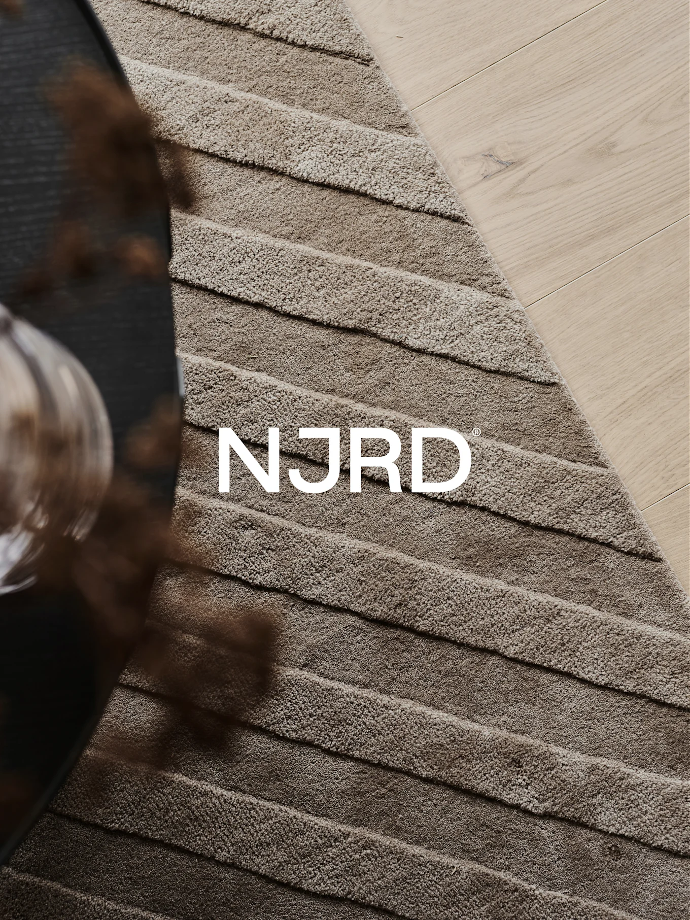 Nærbillede af et lysebrunt luvtæppe med vandrette riller, delvist på et lyst trægulv, med det hvide 'NJRD'-logo.