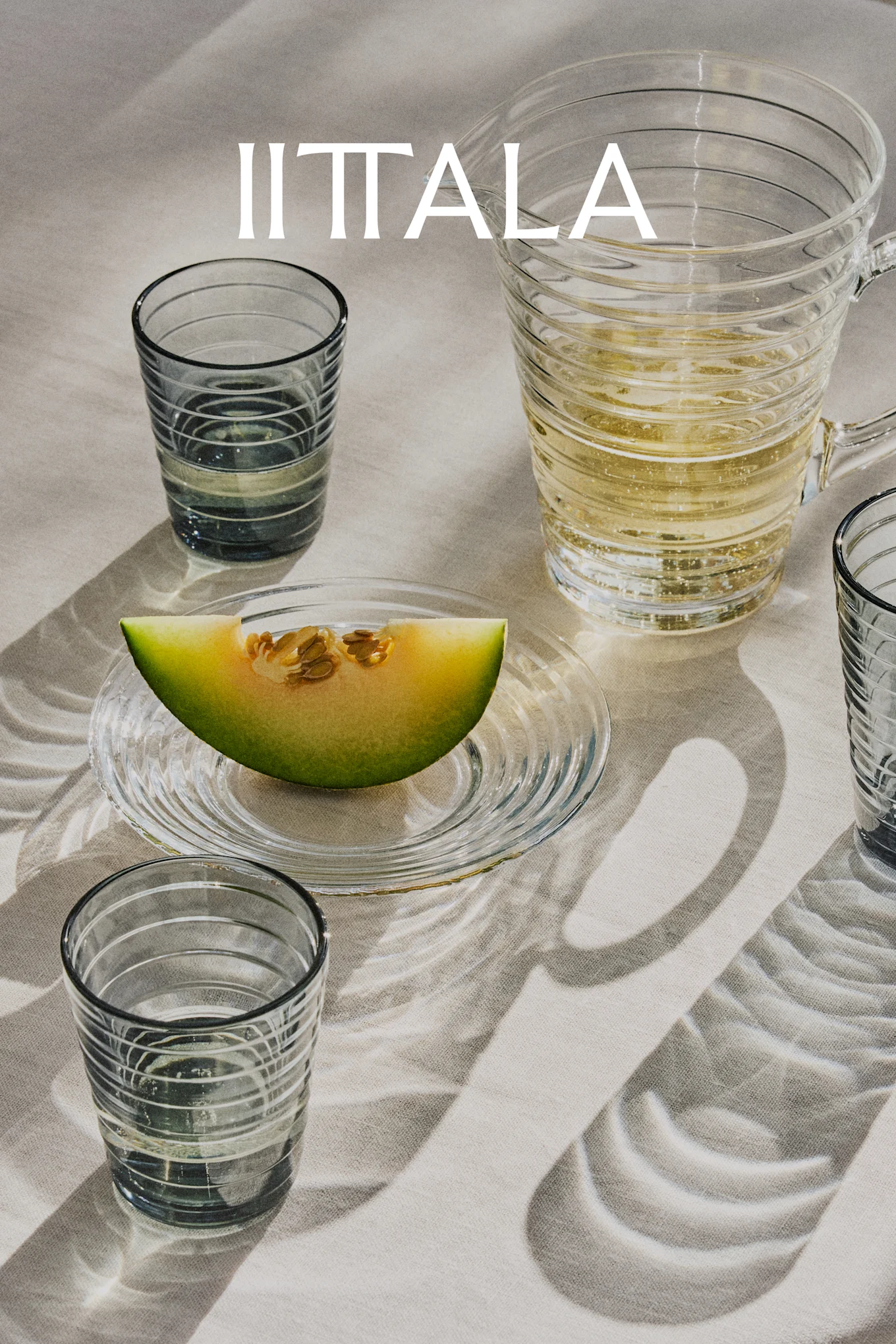 IITTALA logo over glas, en kande med boblende drik og en melonskive på tallerken, på lyst stof.