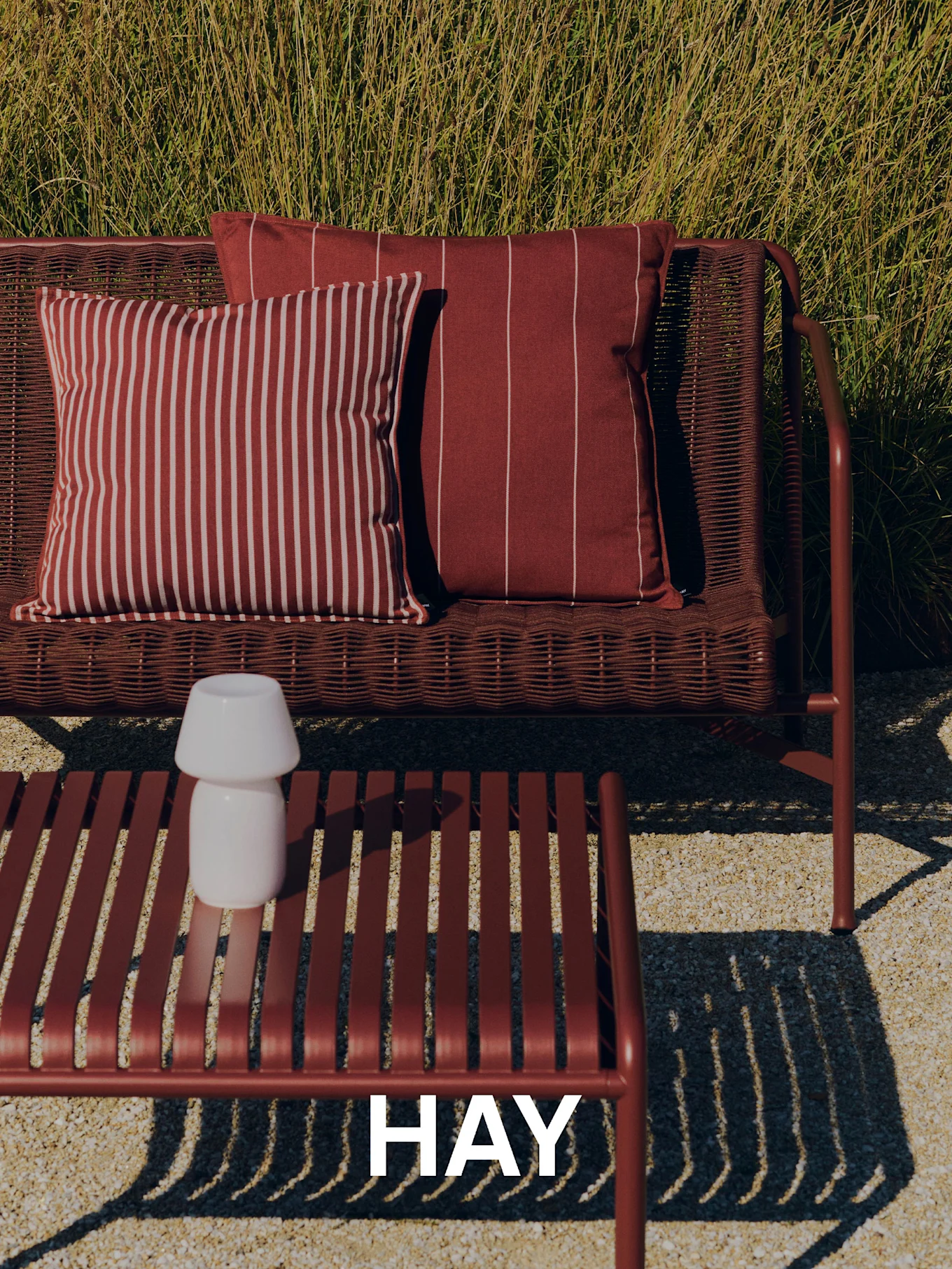 Udendørsscene med en vævet rød sofa, stribede puder, et lamelbord med hvid lampe og højt græs i baggrunden. HAY logo.