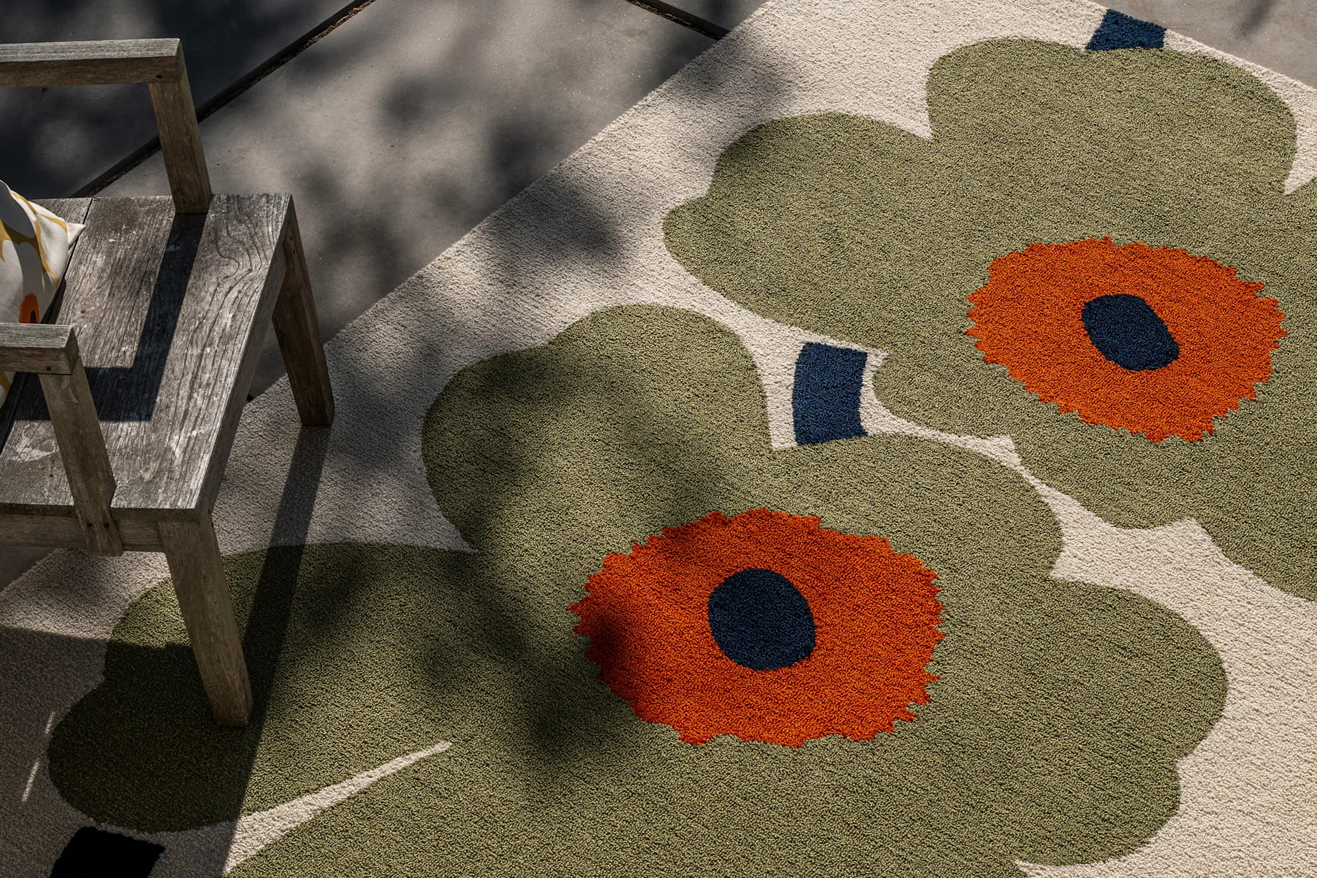 Udendørstæppe med stort oliven grønt, orange og mørkeblåt blomstermønster, ved en rustik træstol i spættet sollys.