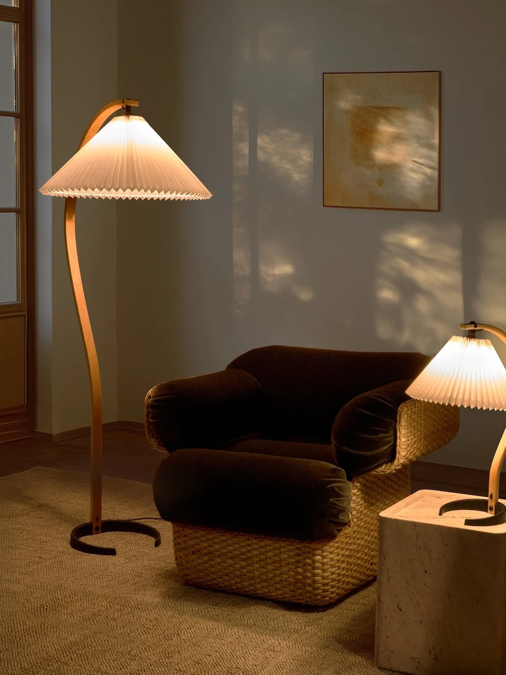 Hyggeligt rum med en buet gulvlampe, mørk lænestol med flettet base og en mindre bordlampe. Varmt lys skaber skygger.
