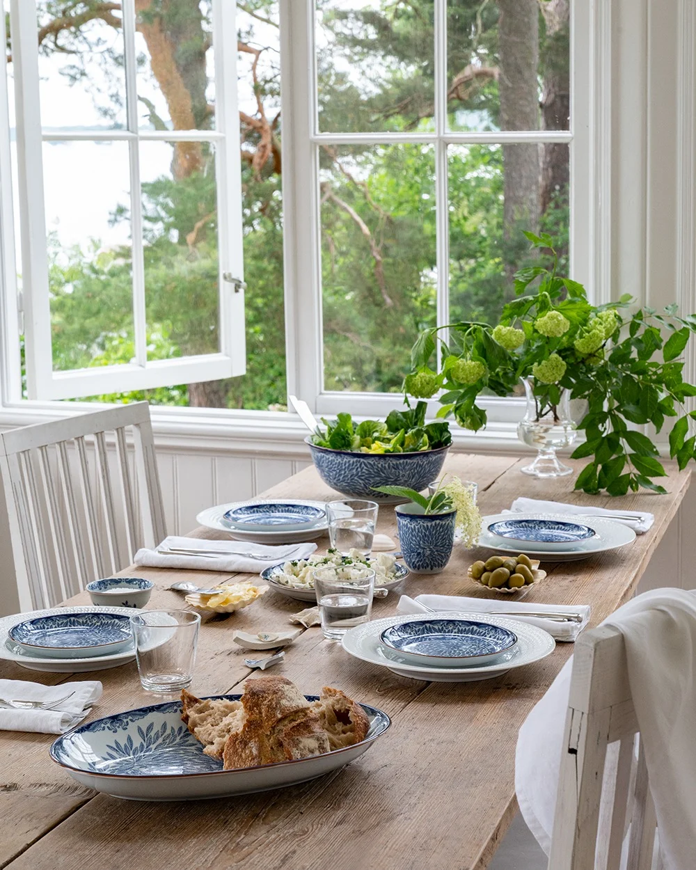 Et rustikt træspisebord dækket med blå og hvide tallerkener, salat, brød og oliven, ved et åbent vindue med udsigt til grønne træer og vand.