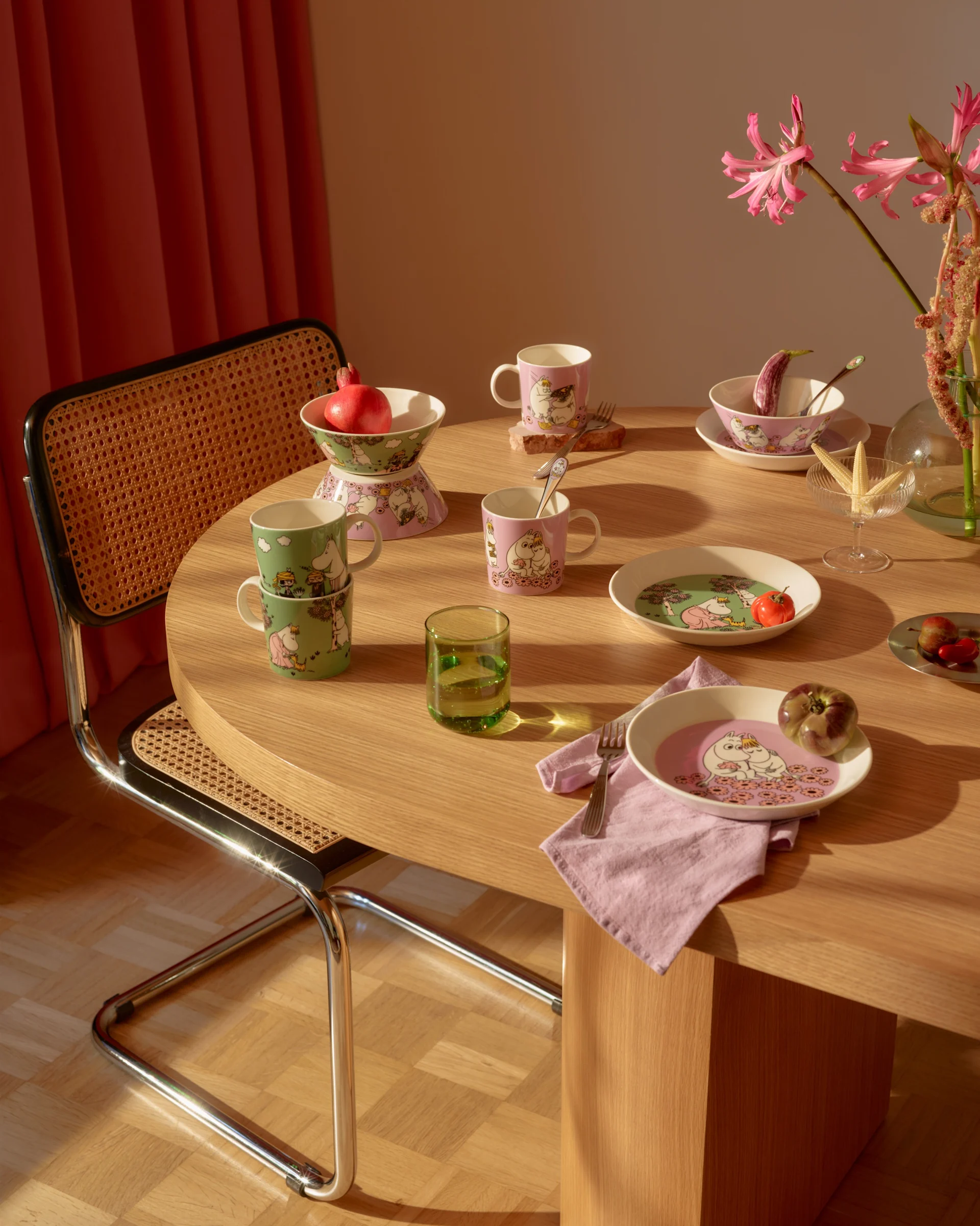 Spisebord med lyserødt og grønt Mumi-stel, friske frugter, et grønt glas og lyserøde blomster.
