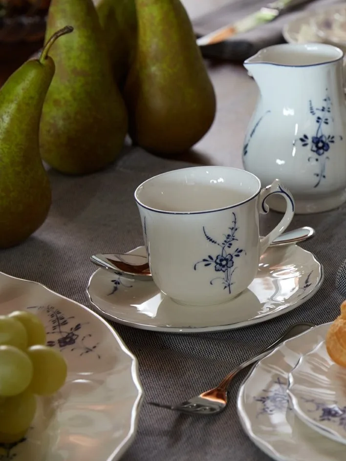 Hvid porcelænsttekop, underkop og kande med blåt blomstermønster på bord med grønne pærer og druer.
