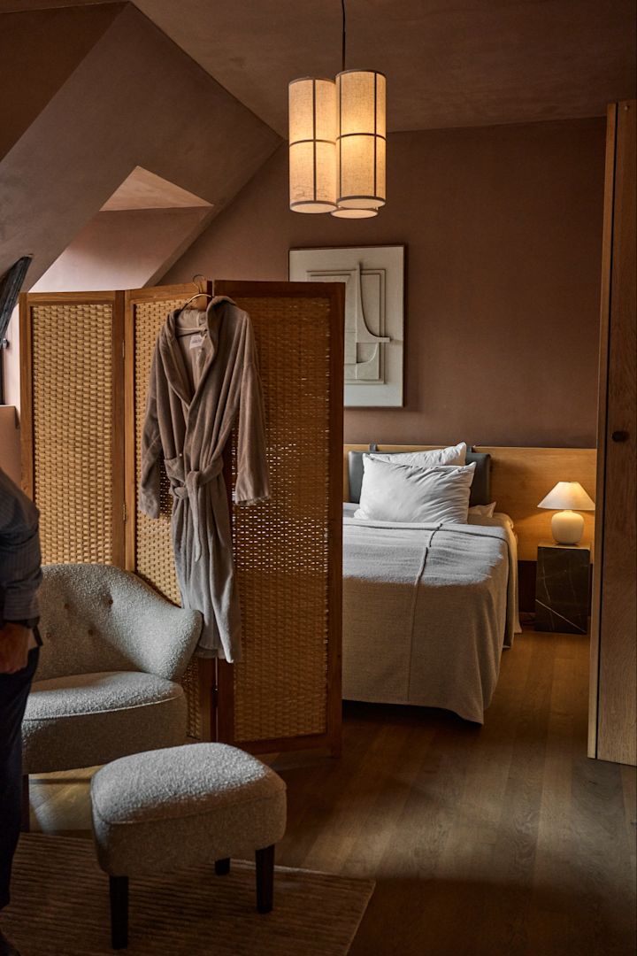Designelskere bør ikke gå glip af en overnatning i Audo House. Her ser du et hyggeligt hotelværelse med en skærmvæg og en morgenkåbe hængende foran en seng. 