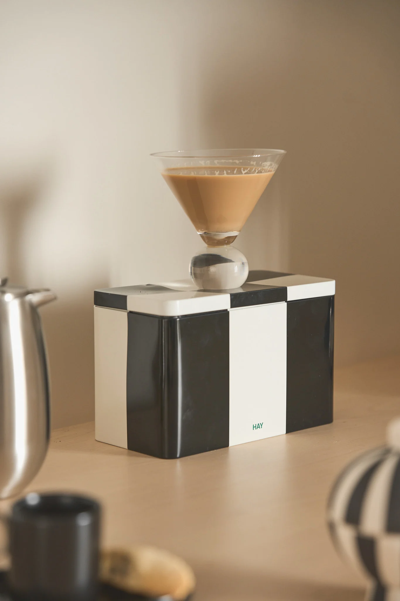 Martini glas med en lysebrun drik og kugleformet stilk på en sort-hvid ternet kasse med 'HAY'-logo.