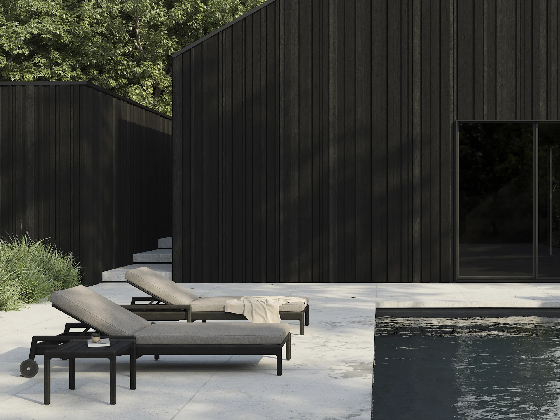 To mørke liggestole med lyse hynder på en terrasse ved en swimmingpool, med moderne mørke træbygninger i baggrunden.