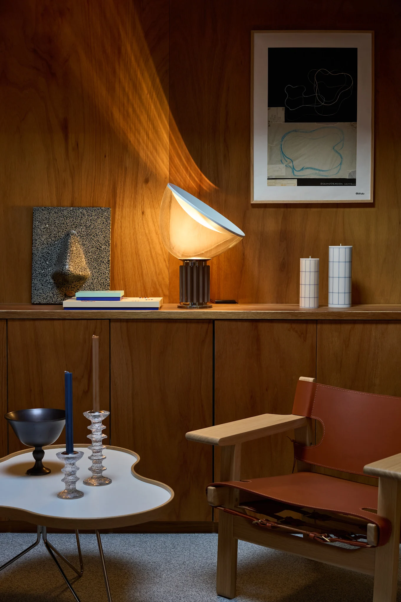 Et opholdsrum med trævægge, en glødende bordlampe, abstrakt vægkunst, dekorative stearinlys, et moderne sidebord og en læderlænestol.