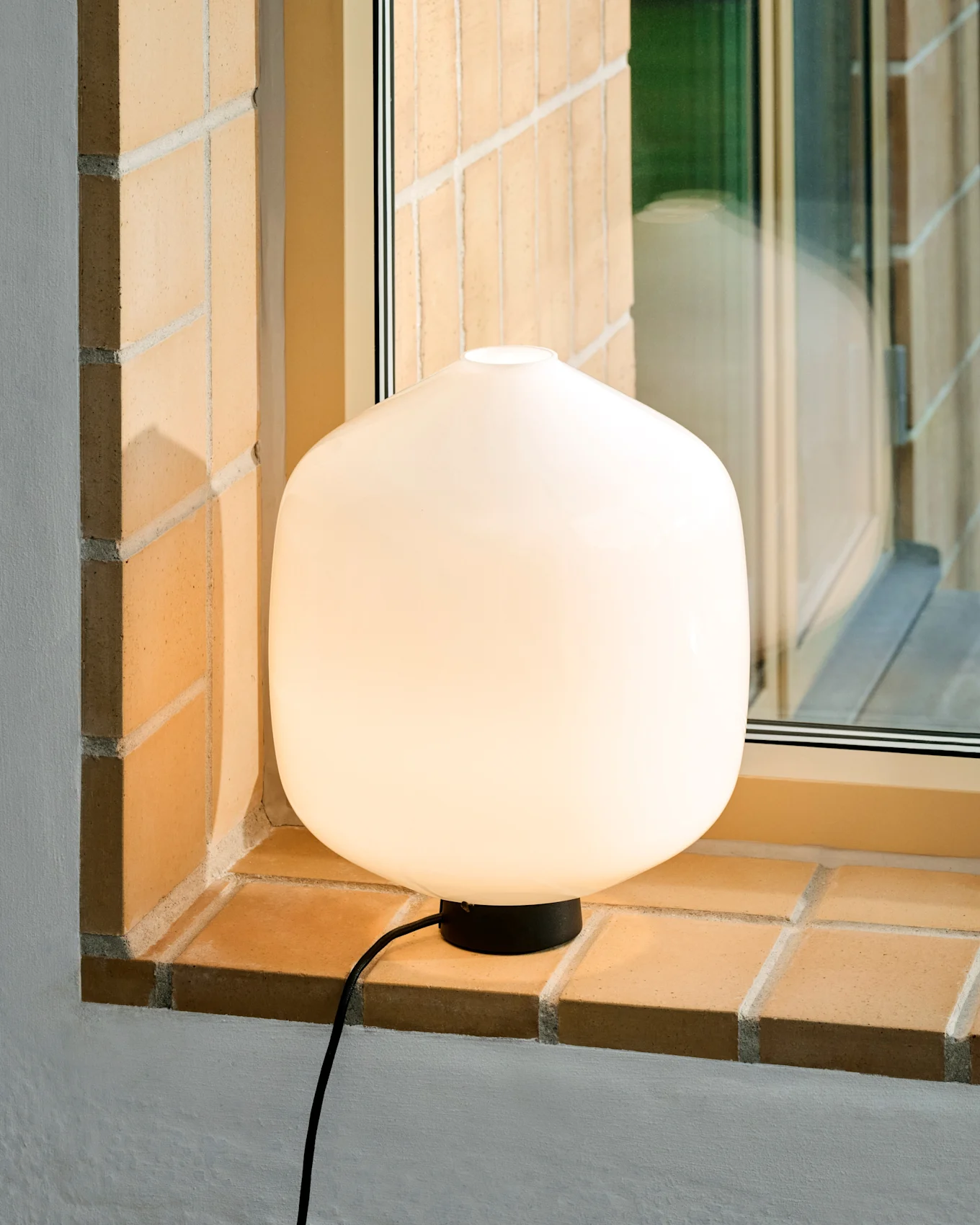 En glødende hvid, afrundet bordlampe med en sort base og ledning sidder på en lys murstensvindueskarm.