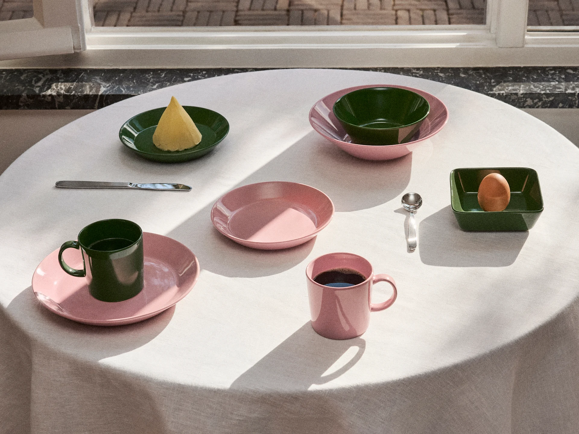 Bord dækket med lyserødt og mørkegrønt service, tallerkener, skåle og krus, med smør, et æg og kaffe i naturligt lys.
