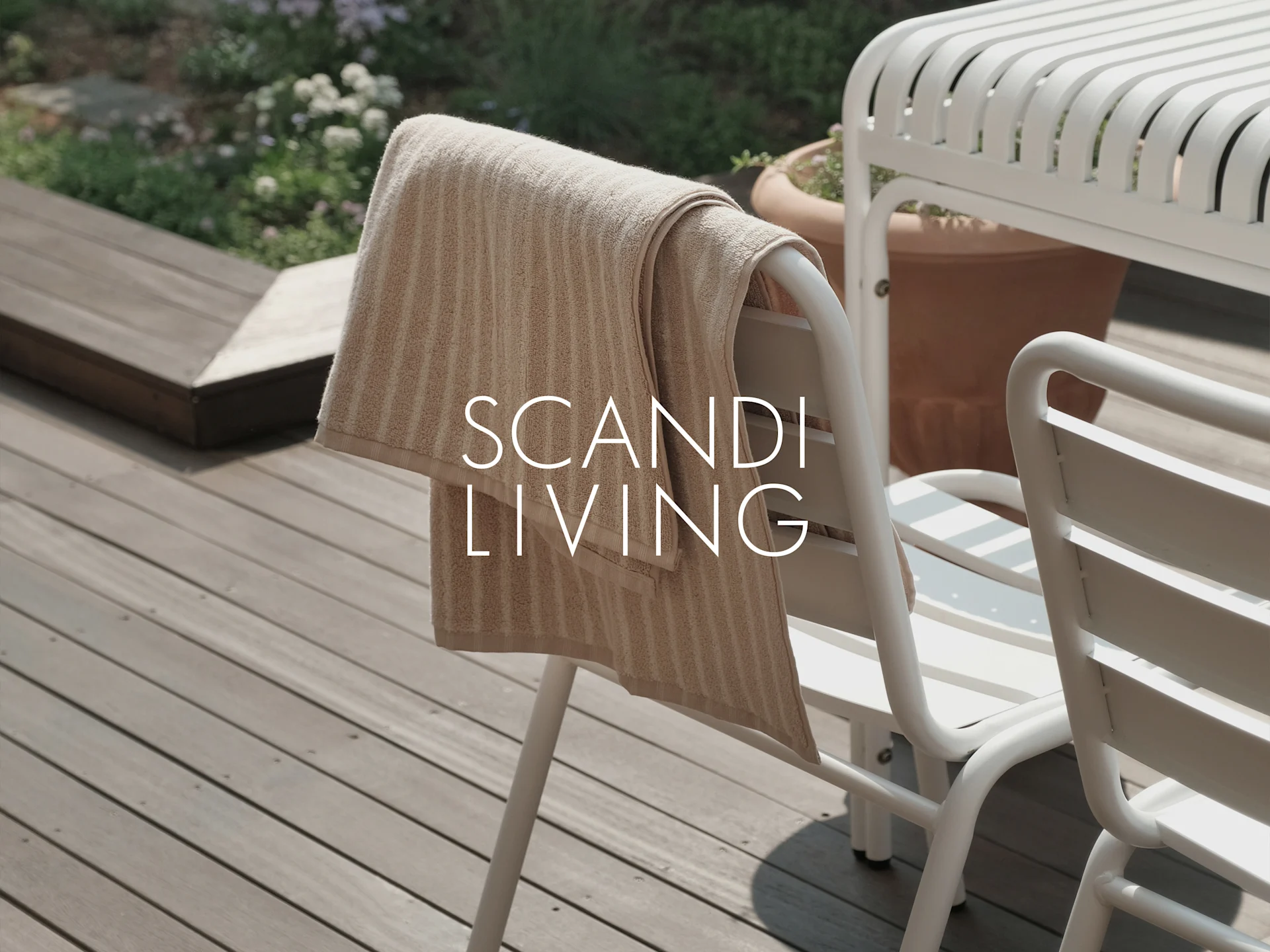 Beige stribet håndklæde på hvid udendørs stol på træterrasse med planter. Tekst: SCANDI LIVING.