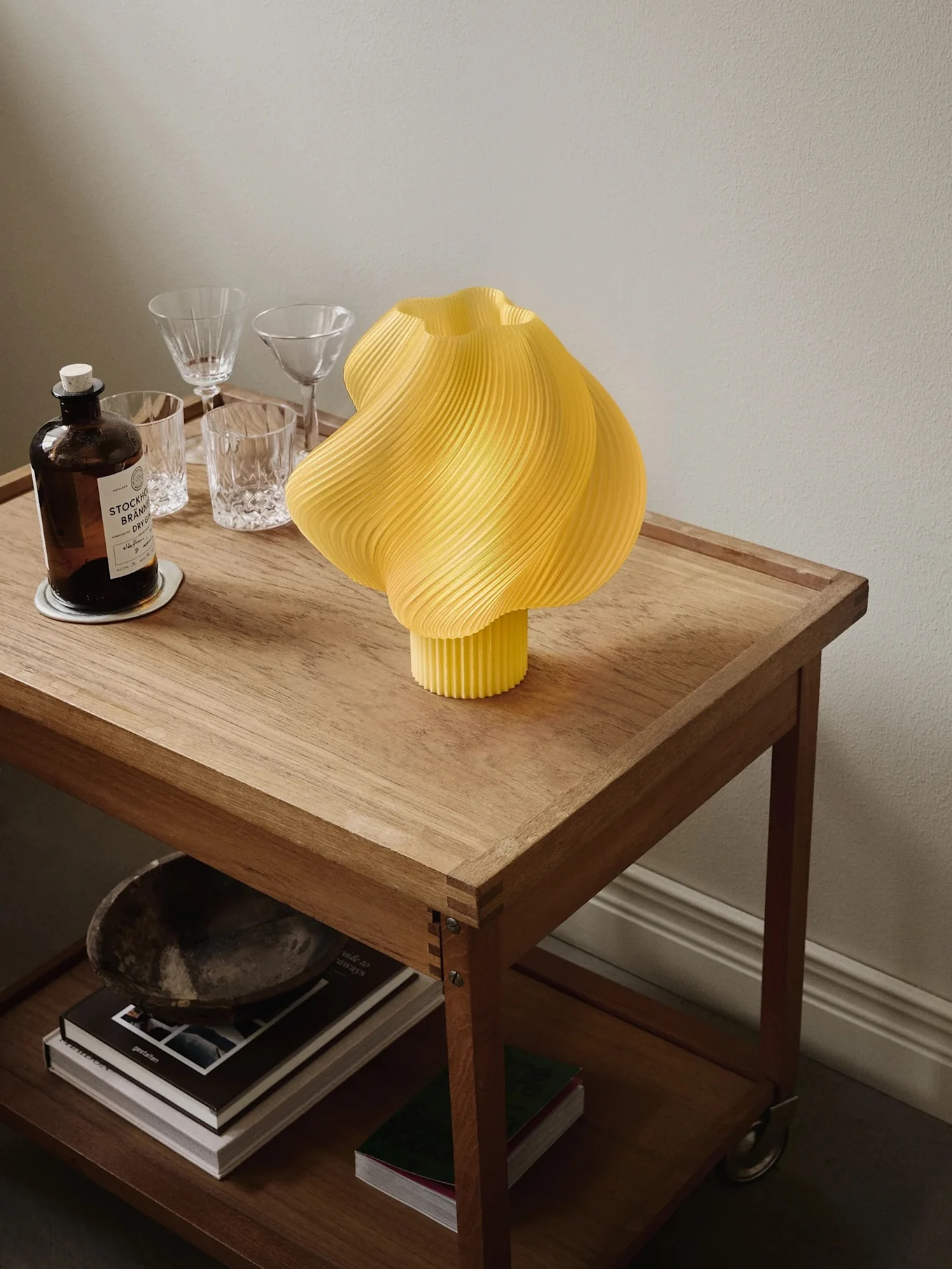Gul plisseret lampe på en rullende trækasse med en ginflaske, glas og bøger på nederste hylde.