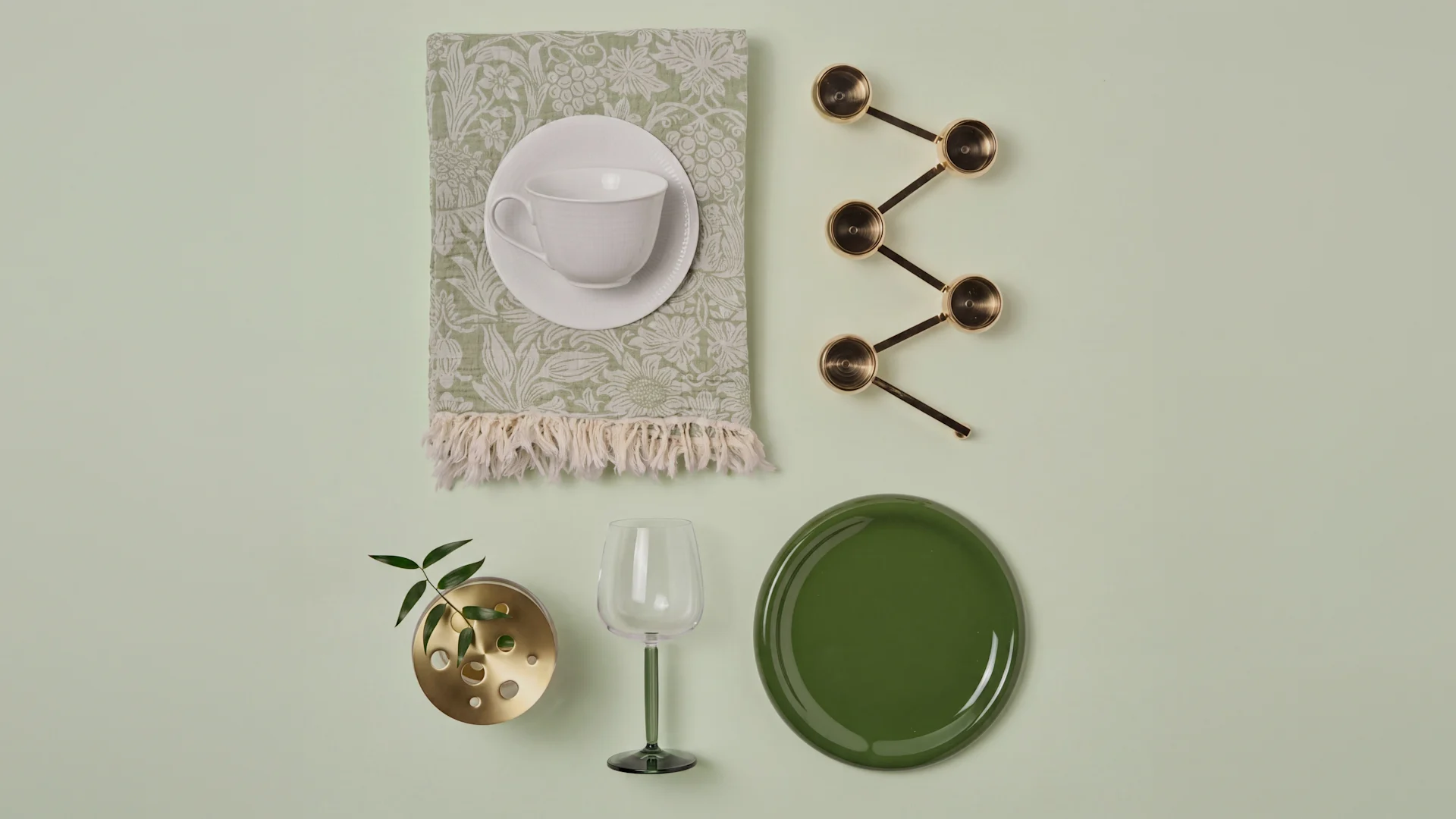 Flad lay: Hvid tekop på grøn serviet, messing måleskeer, messing plantevase, grønt vinglas, mørkegrøn tallerken.