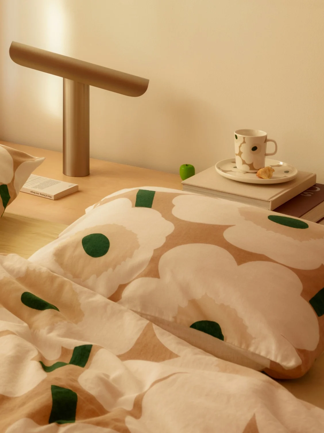 Sengebordsscene med hvidt og beige blomstret sengetøj, en moderne lampe, en kop og en bog på et trænatbord.