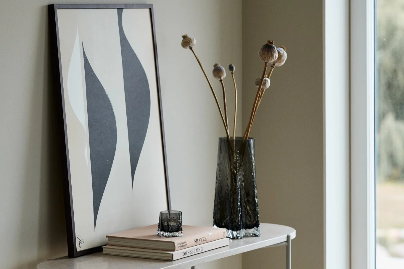 Her kan du se både Gry vasen 30cm og Gry fyrfadsstagen i farven Smoke fra Cooee Design der står på et sidebord foran en plakat.