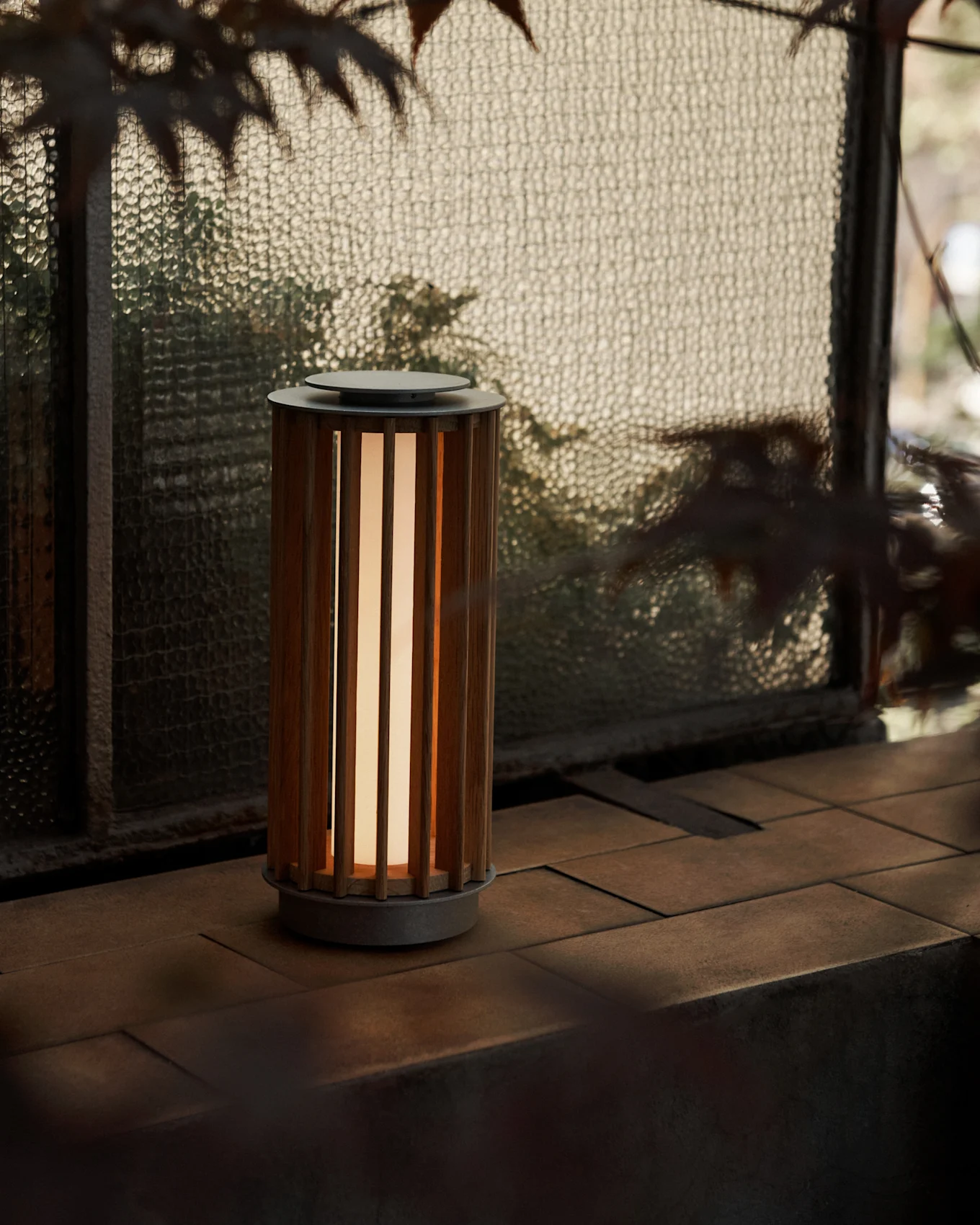 En cylindrisk bærbar lampe med en lameltræskrop og varm glød, på en flisebelagt overflade ved et tekstureret glasvindue.