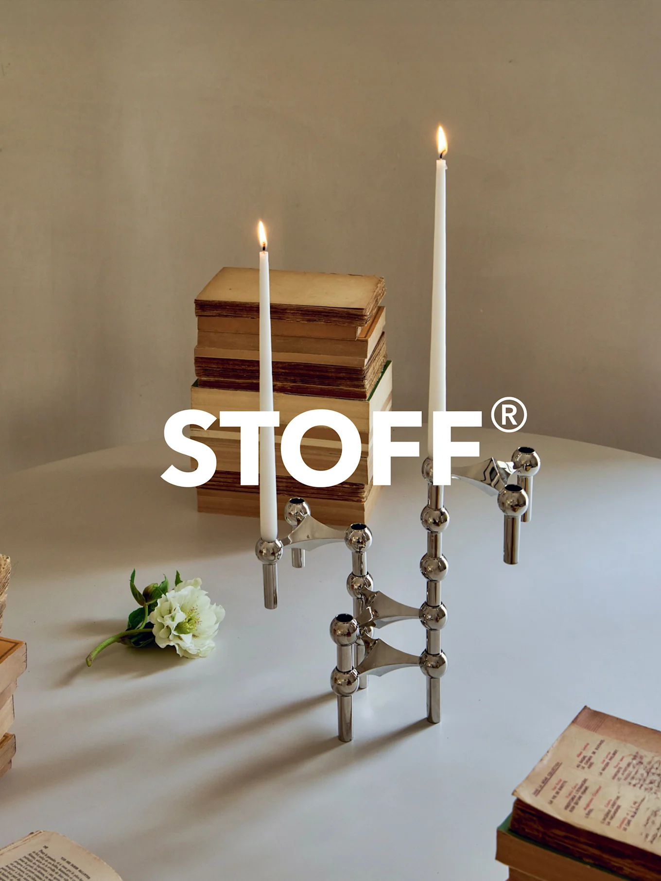 Poleret krom modulær kandelaber med to tændte lys, omgivet af gamle bøger og en hvid blomst på et bord. STOFF® logo.
