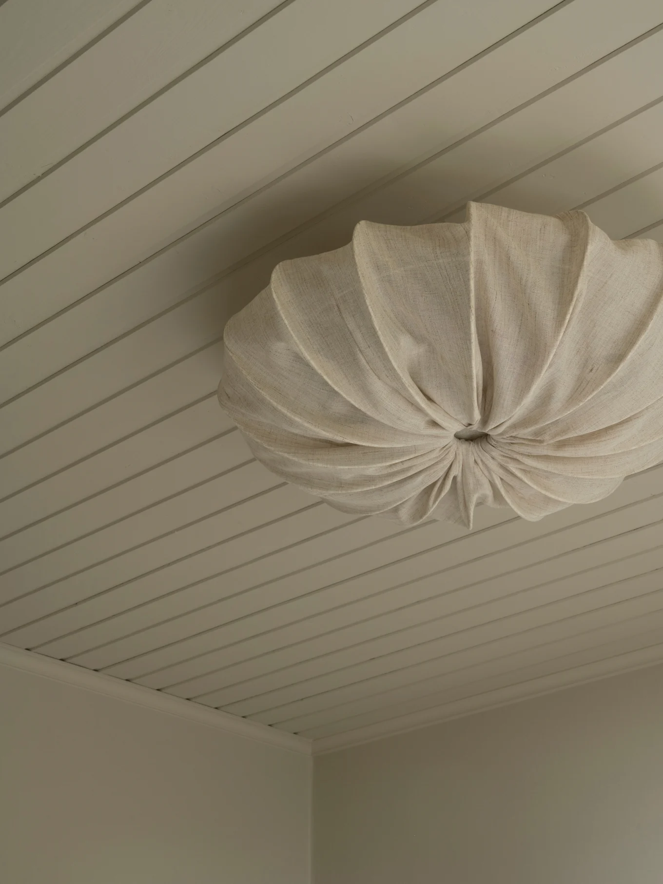 En cremefarvet plisseret loftslampe i stof monteret på et lyst panelloft.