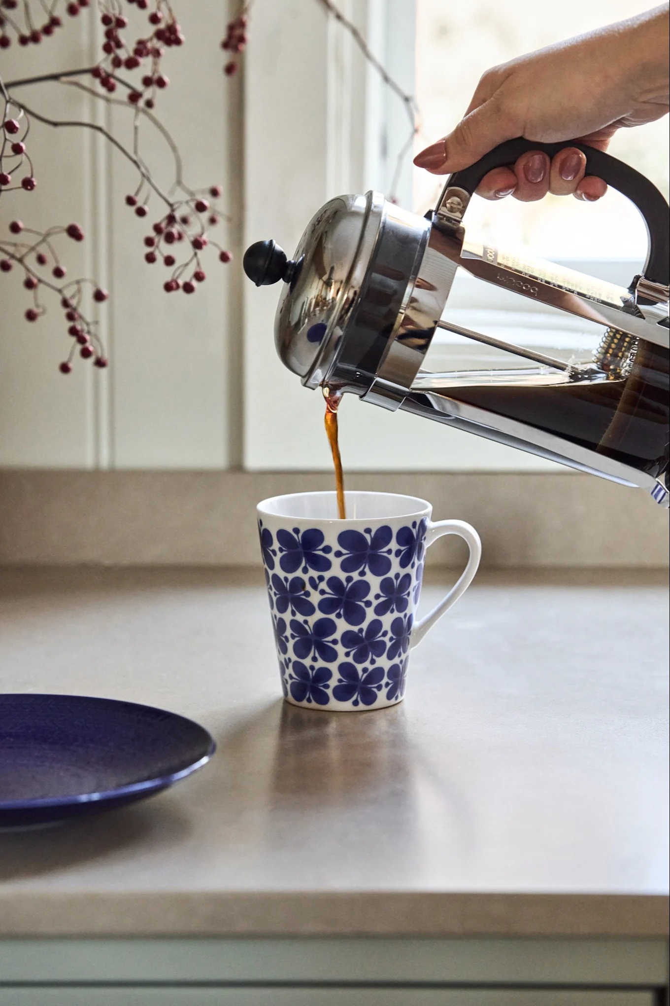 En hånd hælder kaffe fra en stempelkande i et hvidt krus med blåt blomstermønster på en køkkenbænk.