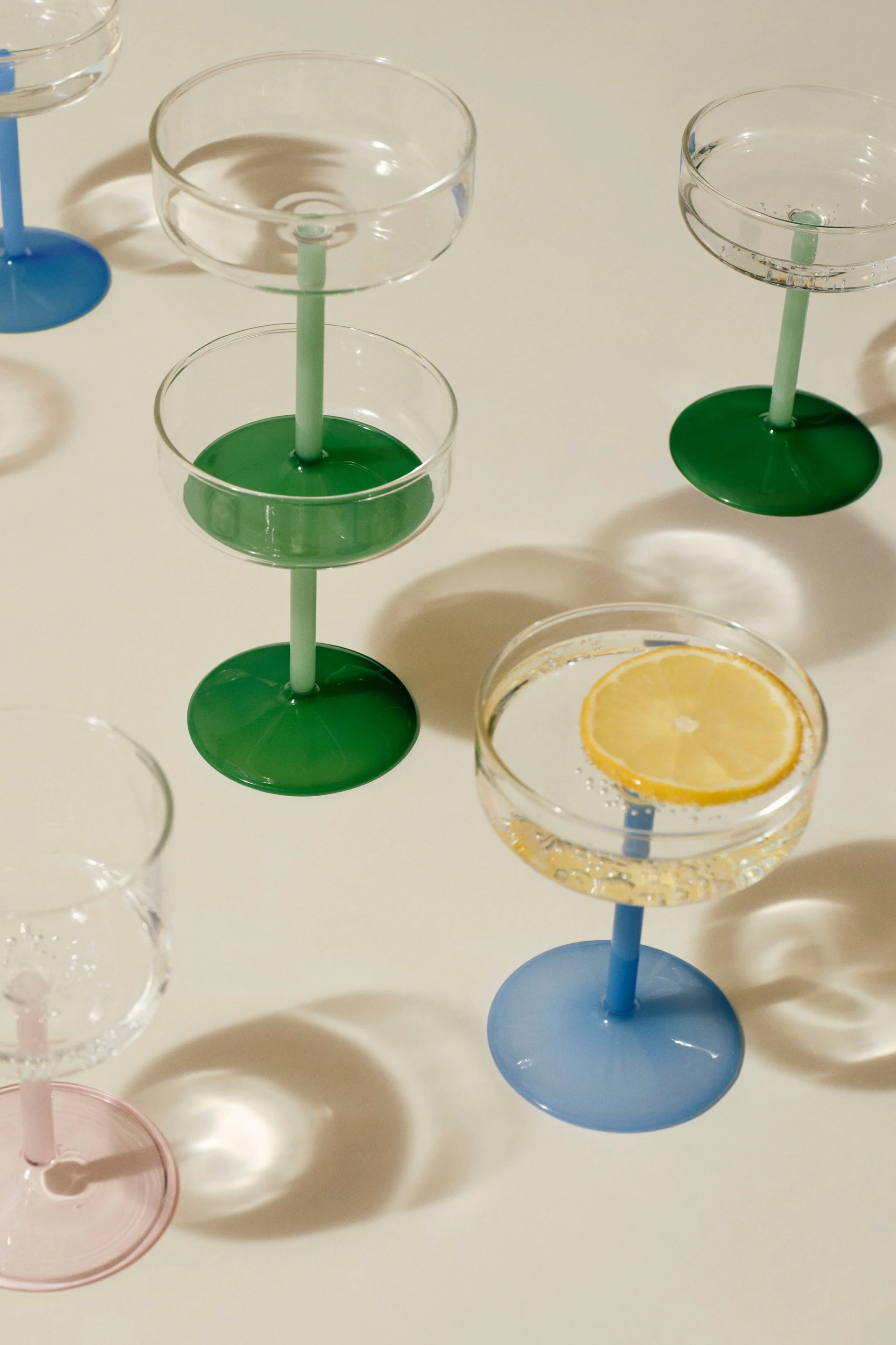 Sortiment af coupeglas med farverige stilke og fødder. Ét glas indeholder en citronskive i danskvand.