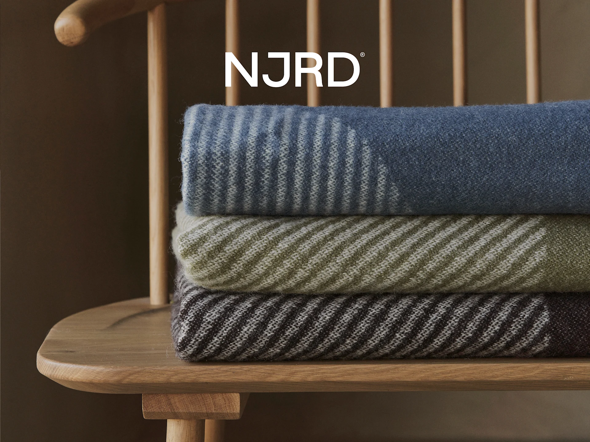 Tre stribede tæpper i blå, oliven og brun, stablet på en træstol med NJRD-logoet over.