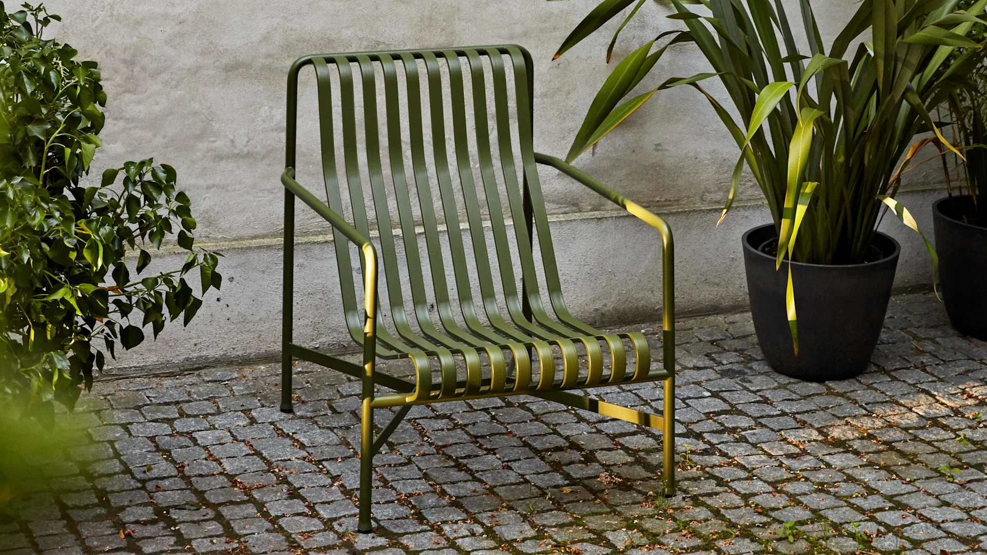 Mørkegrøn udendørs loungestol i metal med lameller på en brostensbelagt terrasse, indrammet af grønne planter.