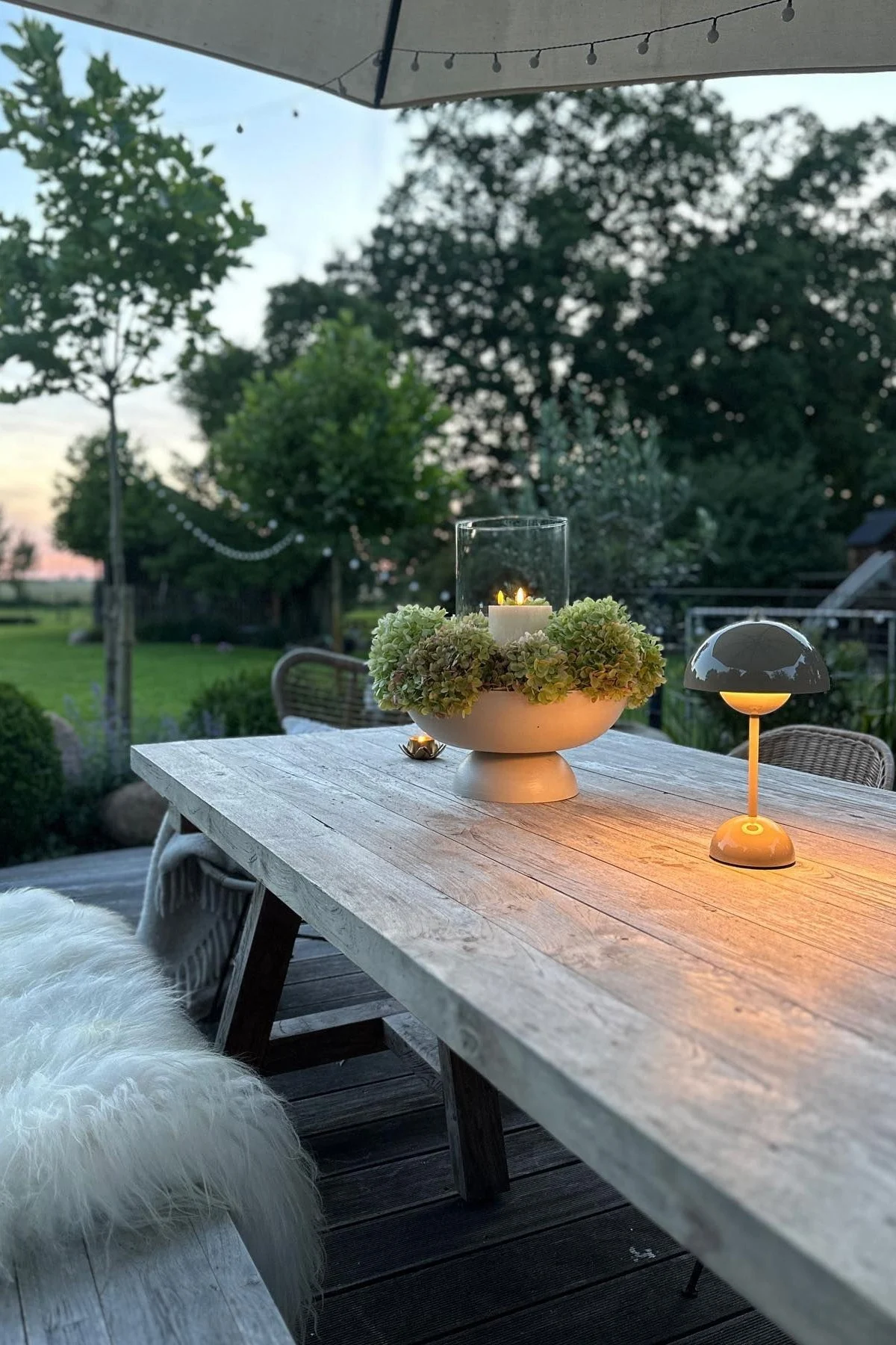 En hyggelig terrasse med et stort spisebord og bænke, dekoreret med lys og en Flowerpot bordlampe fra &Tradition.
