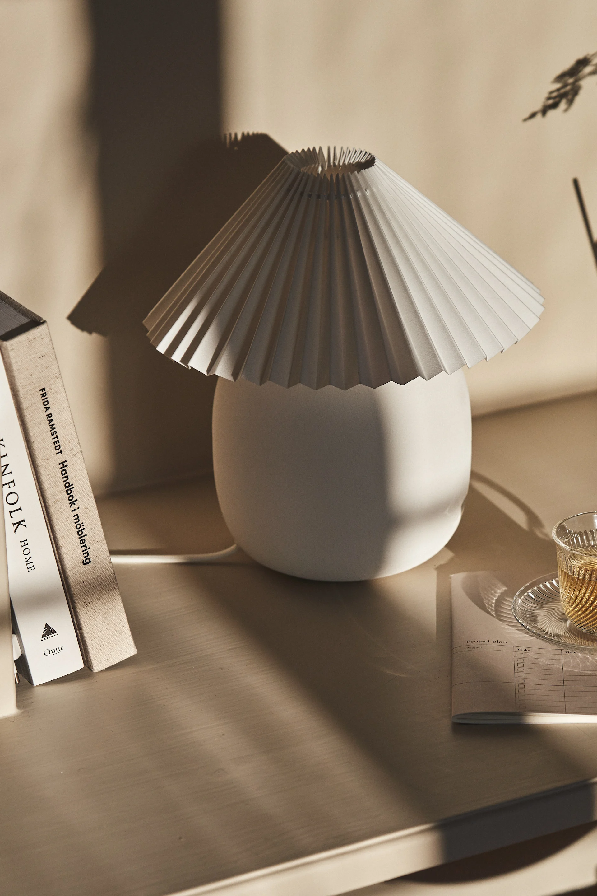 En plisseret hvid bordlampe ved siden af bøger og en kop te på et lyst træskrivebord, med bløde skygger.