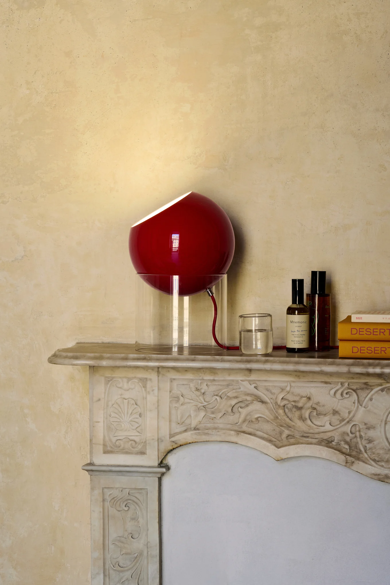 En rød kugleformet lampe med gennemsigtig fod står på en udsmykket marmorkaminhylde ved siden af et glas vand, to flasker og to gule bøger.