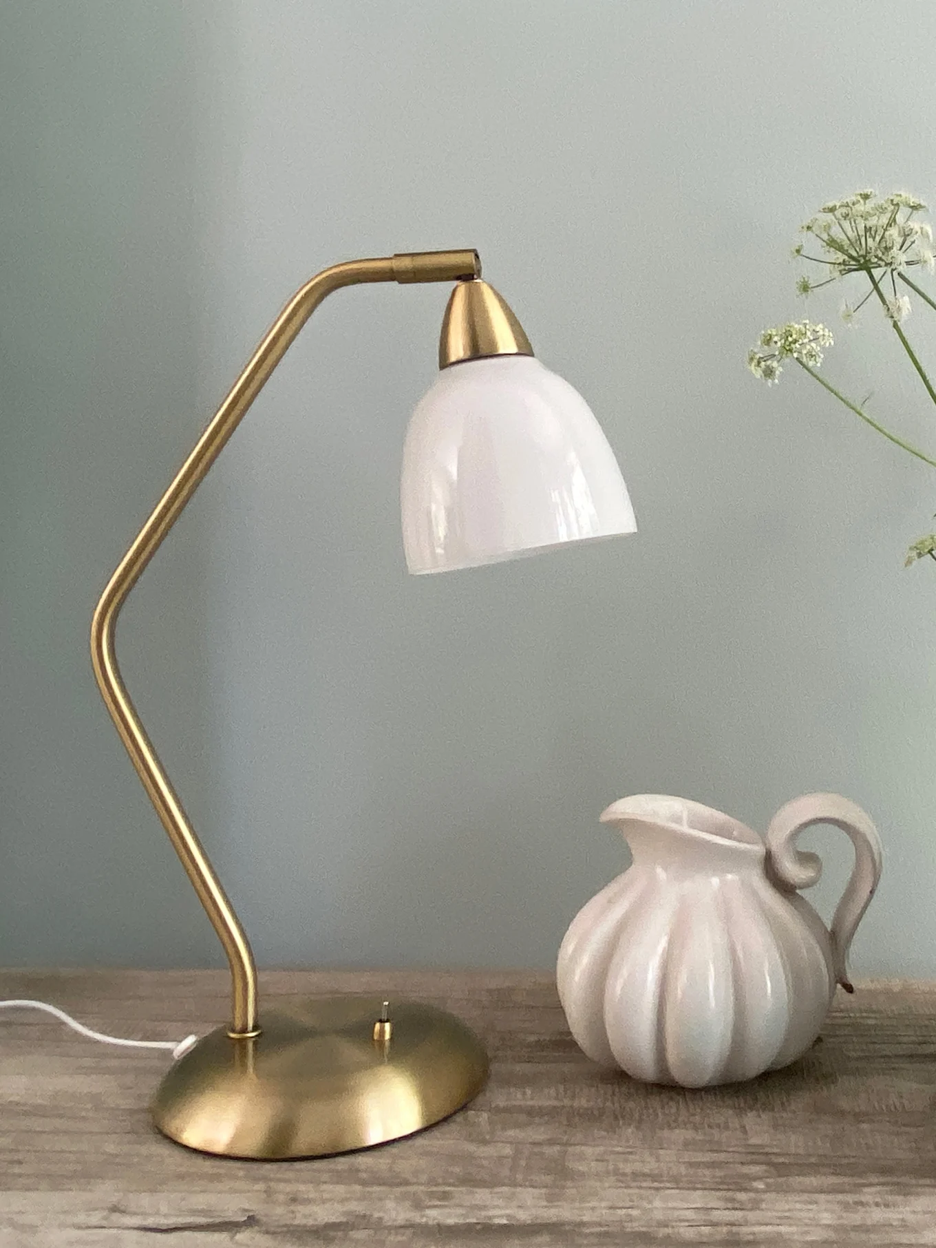 En messing bordlampe med en hvid glasskærm står ved siden af en hvid riflet keramik kande på en træoverflade.
