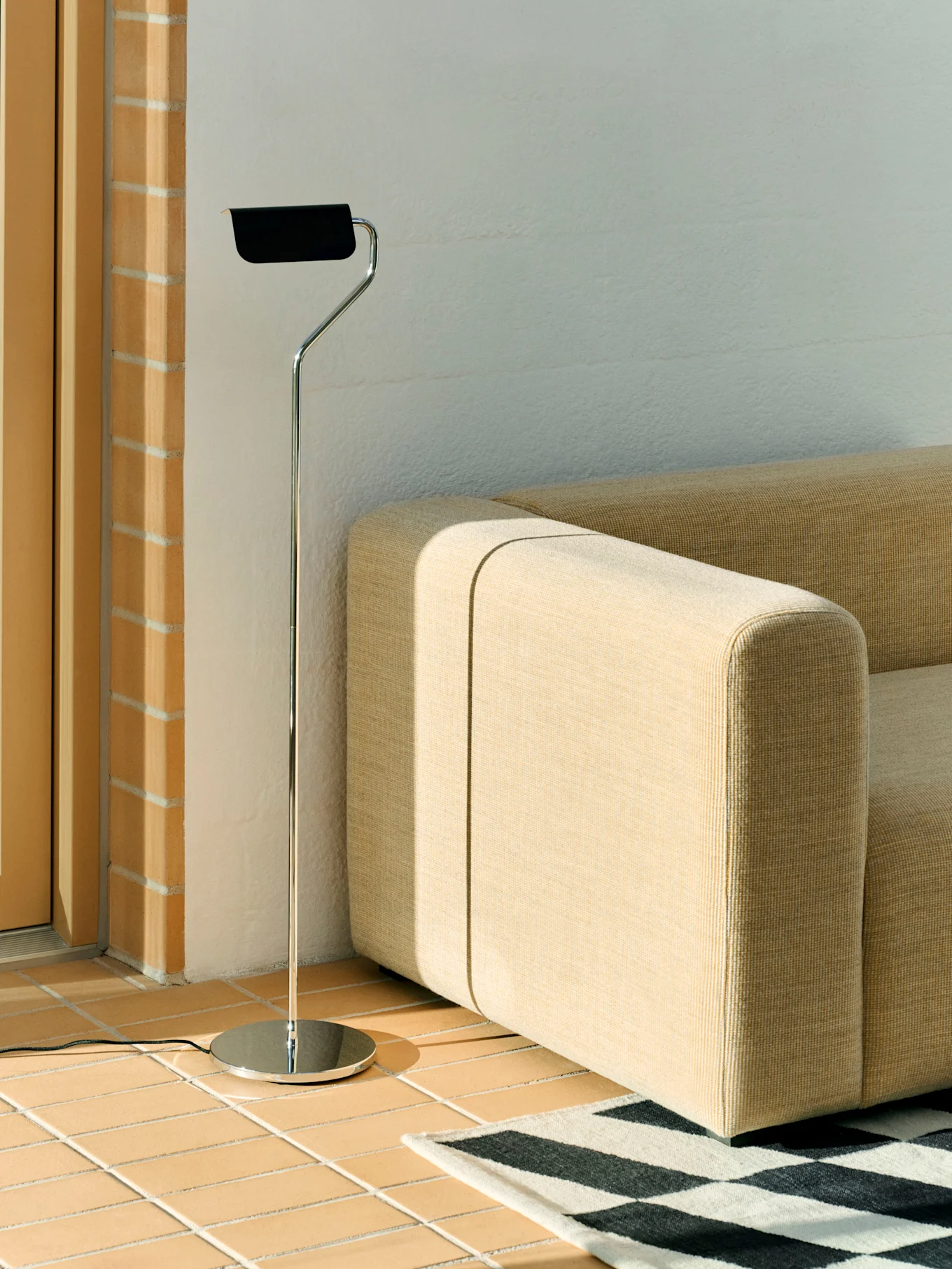 En kromgulvlampe med sort skærm står ved siden af en beige modulsofa og et sort-hvidt geometrisk tæppe på et flisebelagt gulv.