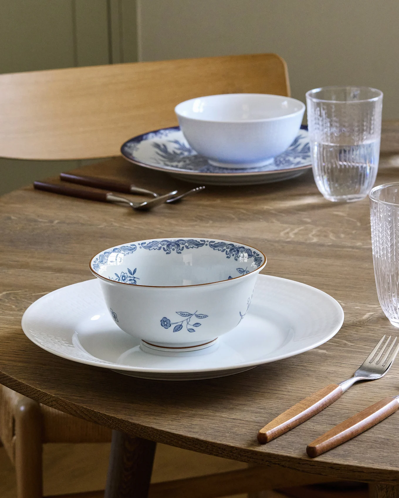 Spisebord dækket til to med hvide og blåmønstrede skåle på hvide tallerkener, bestik med træskaft og vandglas.