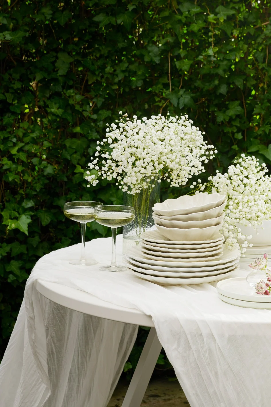 Udendørs bord dækket med hvidt service, champagneglas og brudeslørblomster mod grønt løv.